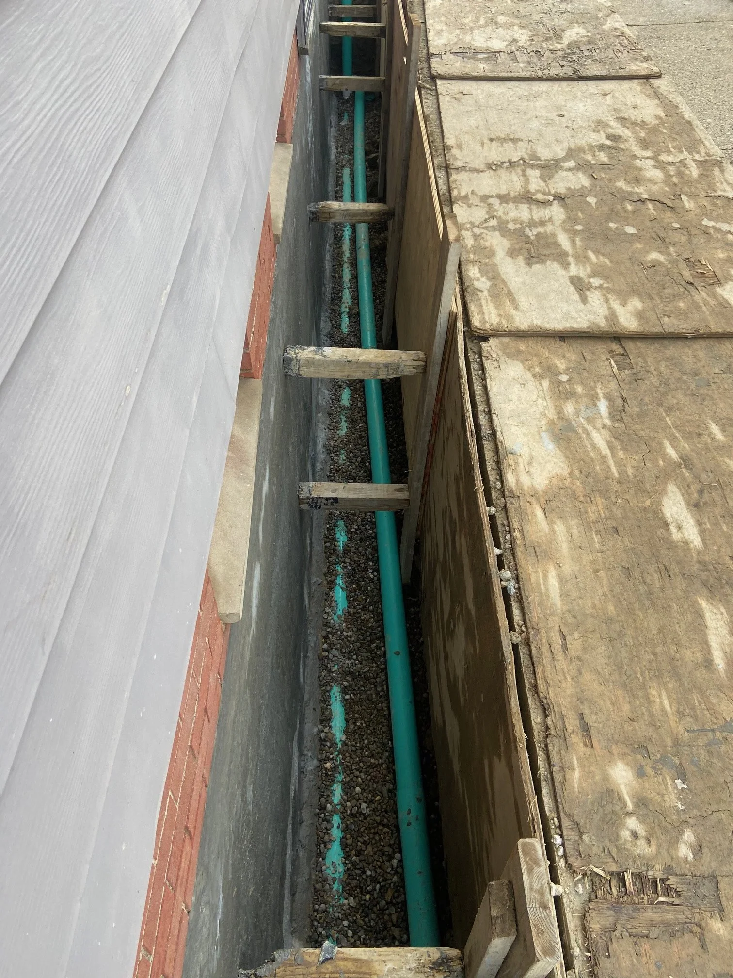 Construction waterproofing site showing a trench with a green PVC pipe installed, surrounded by wood framing and temporary protective panels.