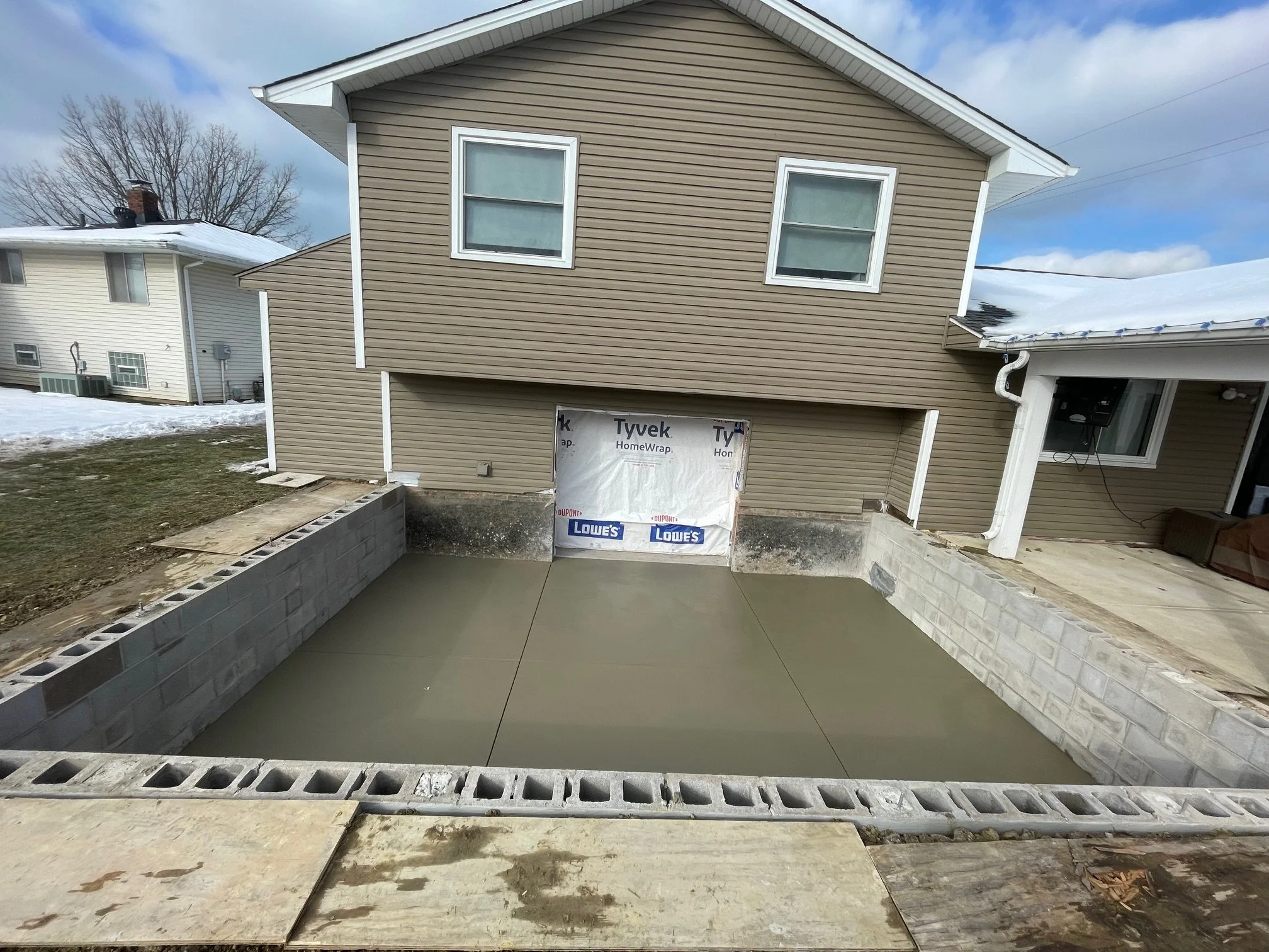 A house with a garage foundation poured and concrete slab set, surrounded by cinder block walls, in a yard with patches of snow and construction materials nearby.