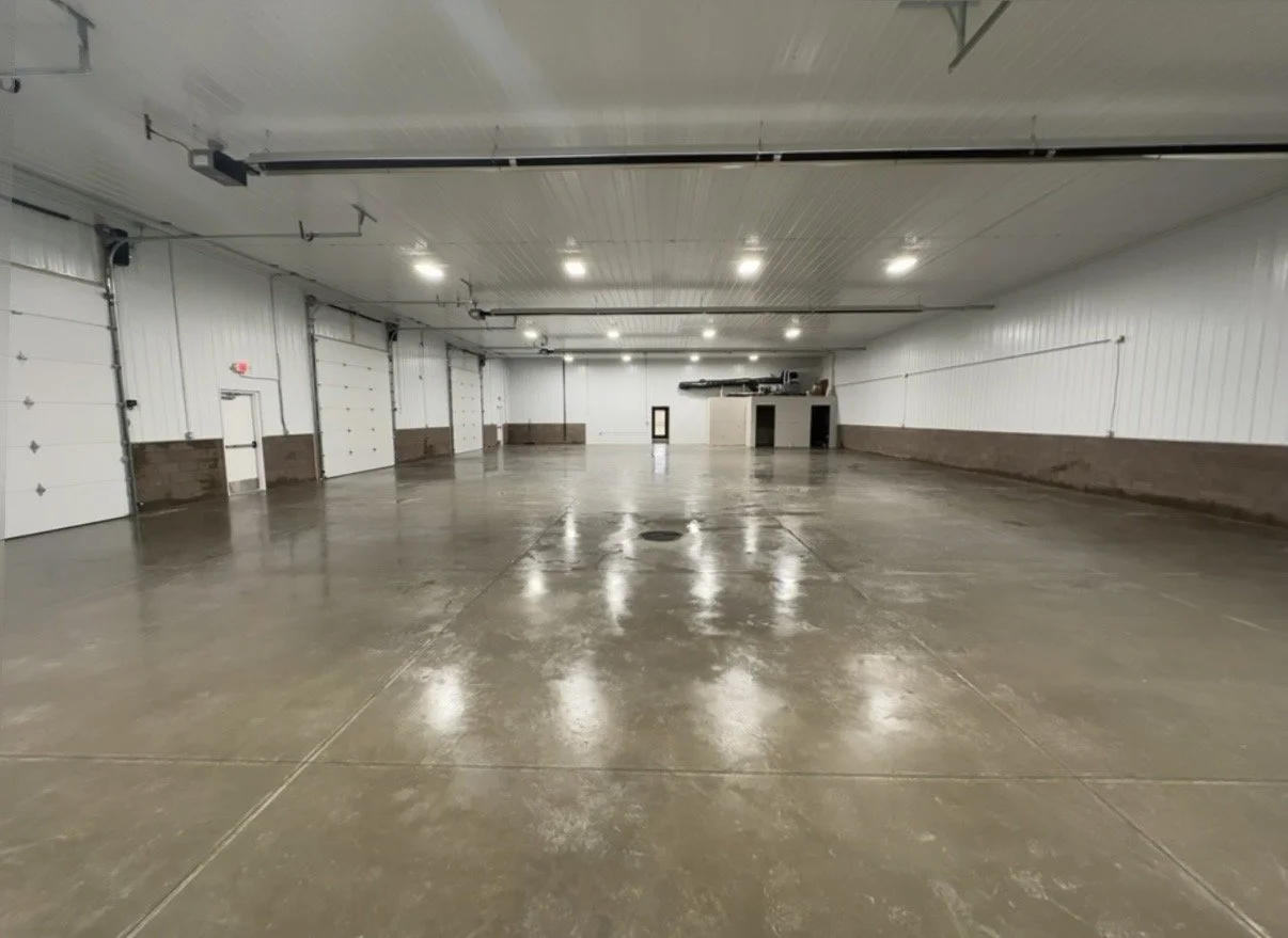 Empty indoor warehouse or garage with polished concrete floors, white panel walls, and several large closed garage doors.