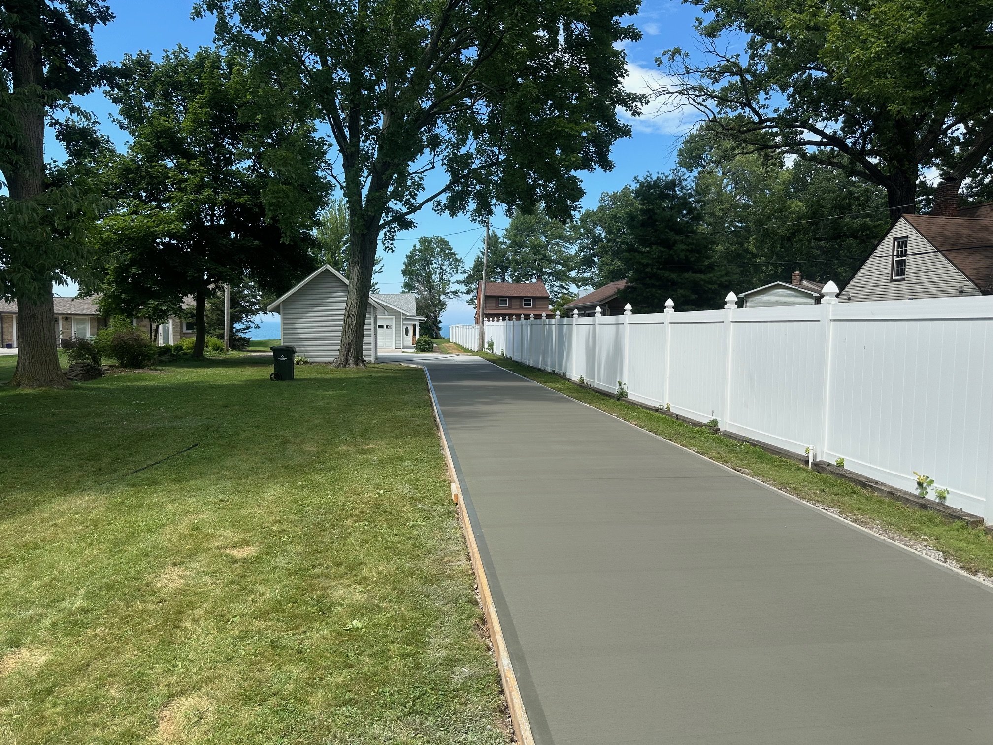 A suburban backyard with a freshly poured concrete sidewalk next to a white privacy fence, green grass, mature trees, and neighboring houses under a partly cloudy sky.