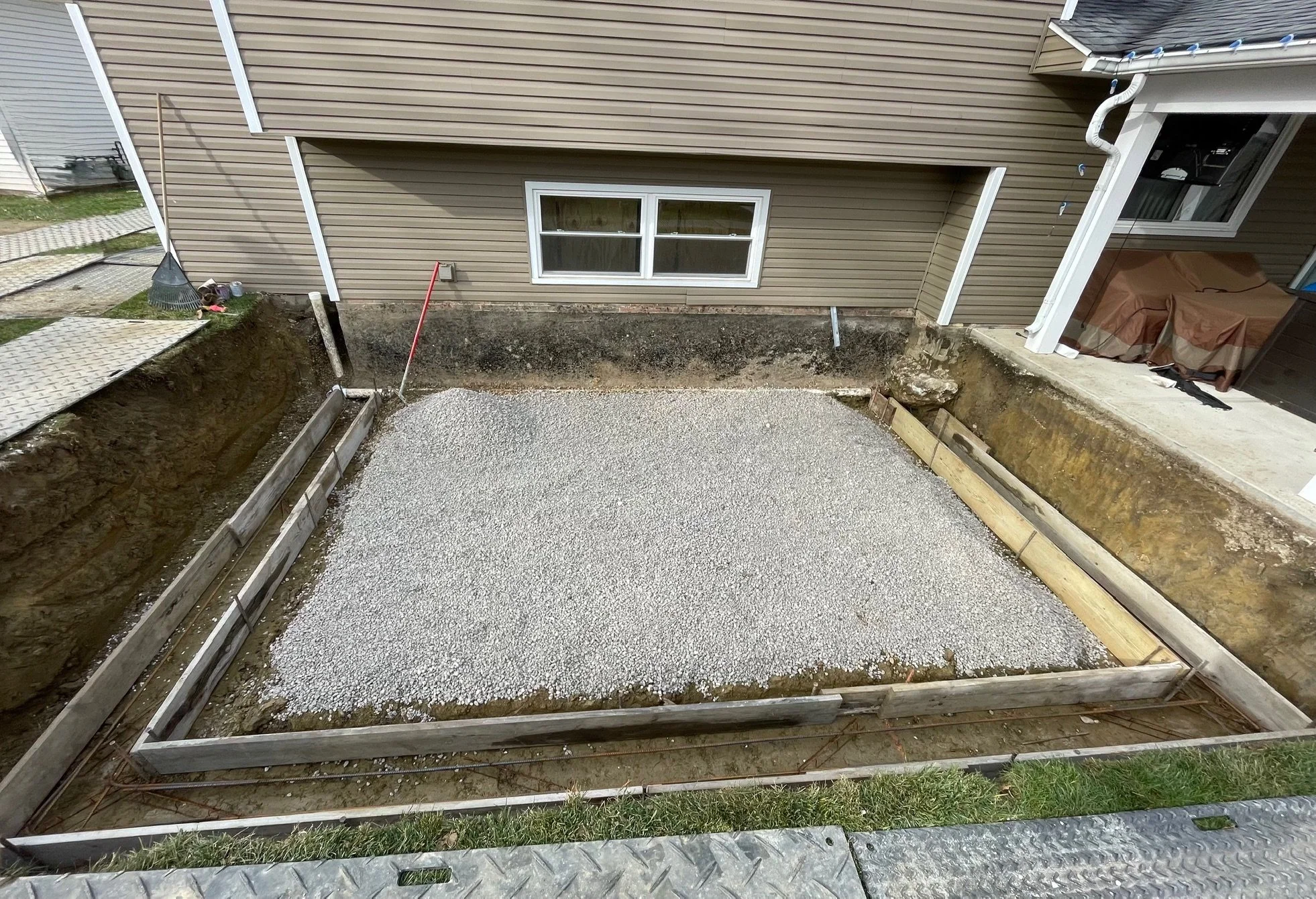 A backyard with a framed rectangular area filled with gravel, surrounding a house with beige siding. There are stairs leading to a side entrance and some tools and construction materials nearby, indicating ongoing landscaping or foundation work.