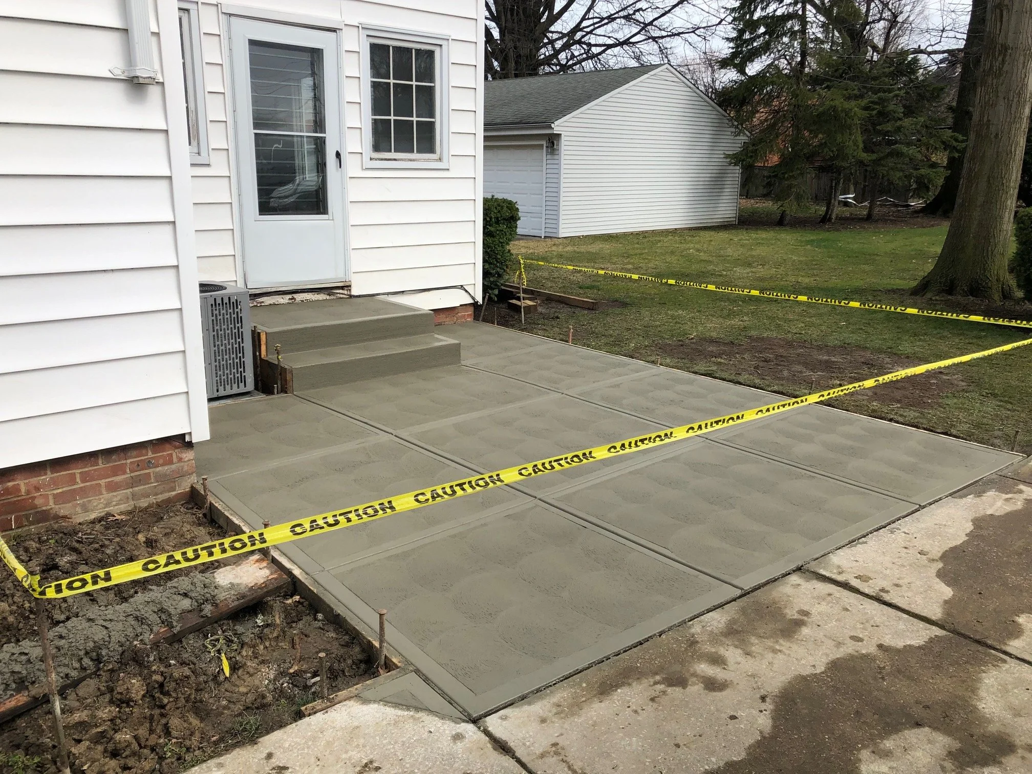 Freshly poured concrete sidewalk with steps leading to a house door, surrounded by caution tape indicating ongoing construction.