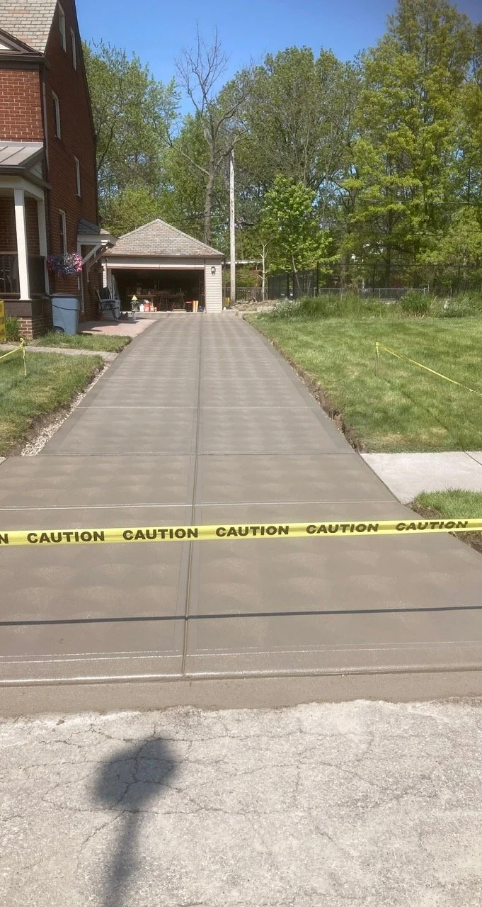 Concrete driveway leading to a garage, cordoned off with yellow caution tape, in a residential area with grass and trees.