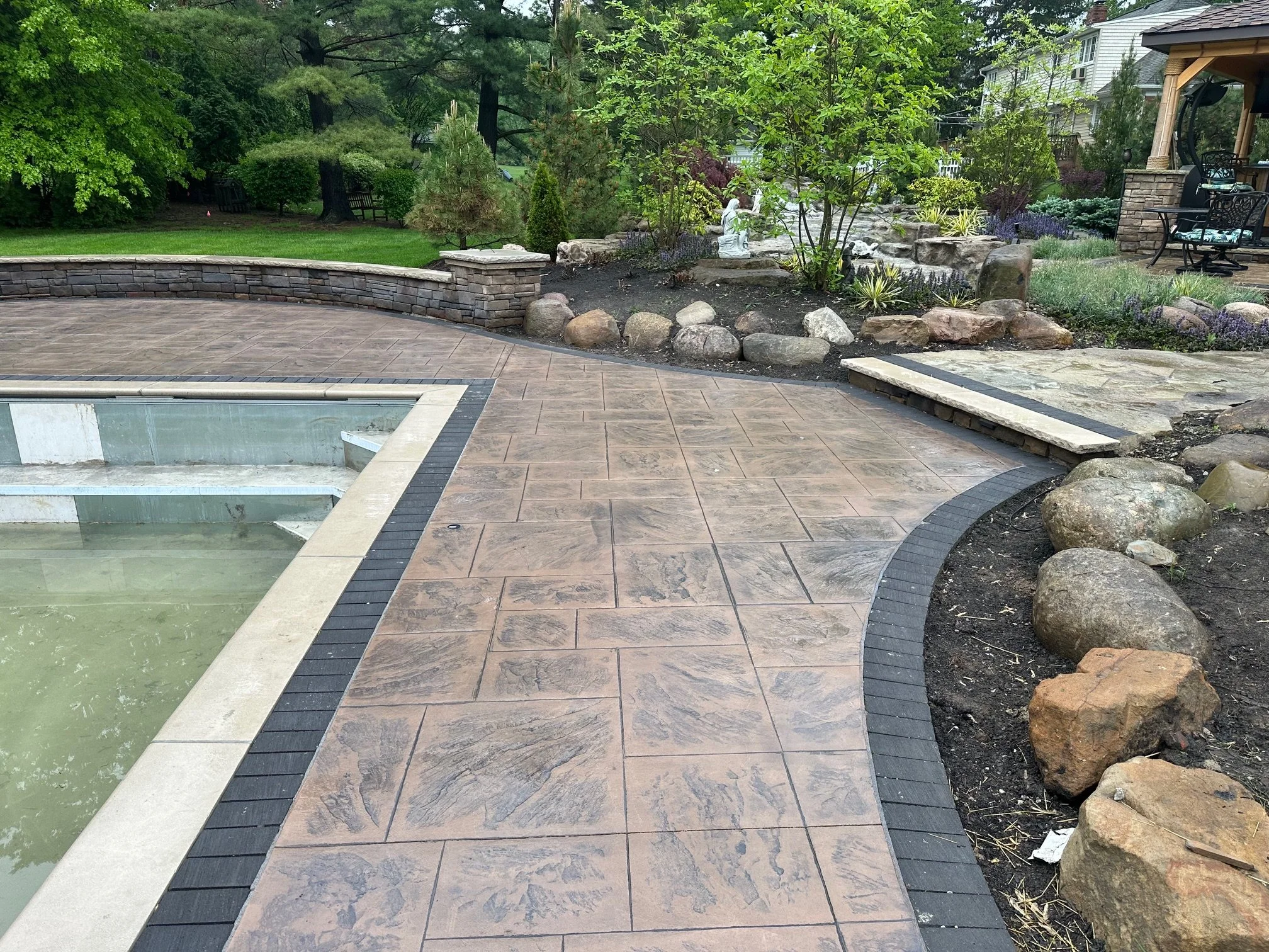 A backyard patio with a water feature, surrounded by a well-maintained garden with potted plants, trees, and bushes, and a house with outdoor furniture on a porch in the background.