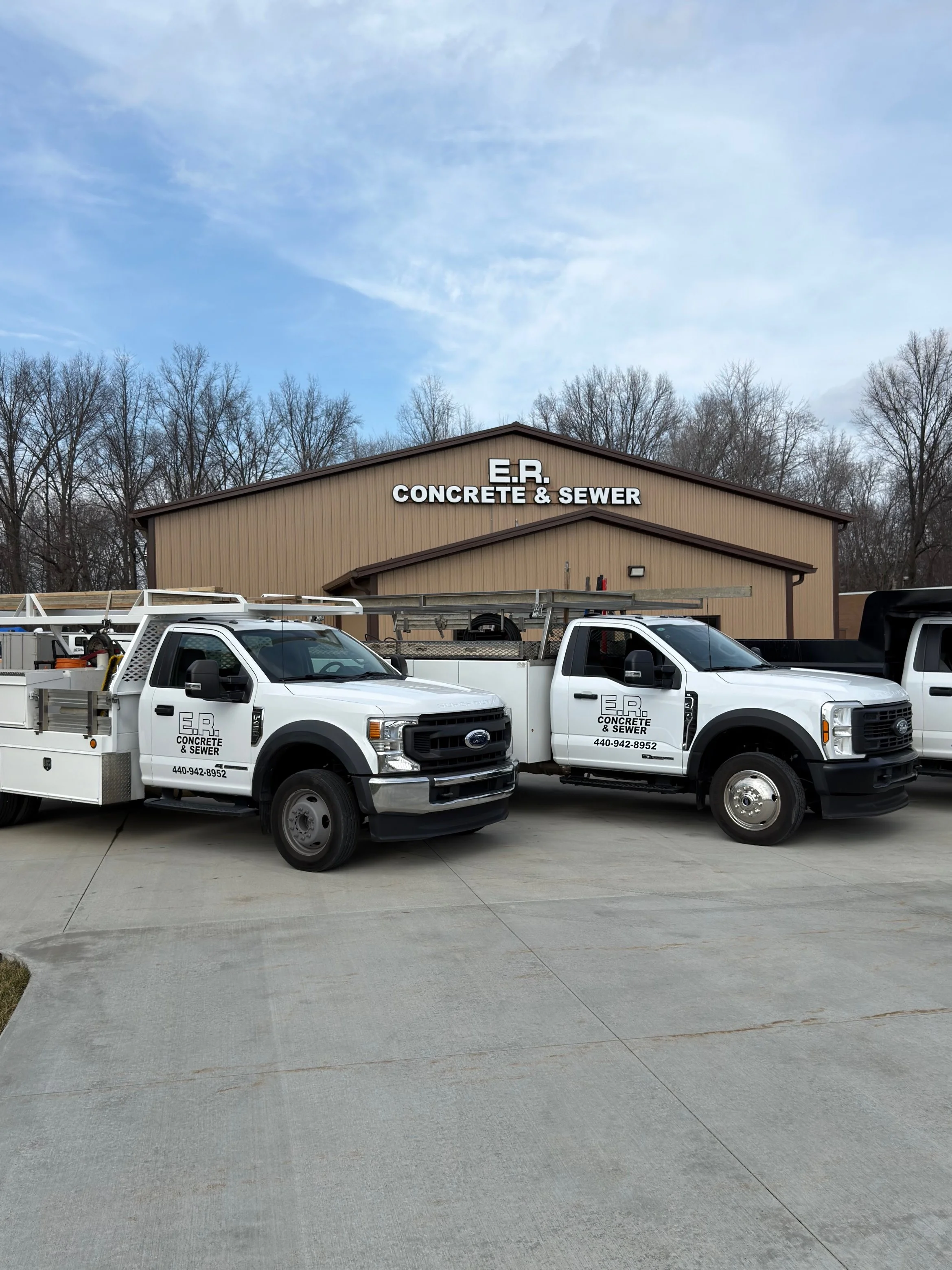Two white utility trucks parked in front of a beige building with a sign that reads 'E.R. CONCRETE & SEWER.' The trucks have company branding and phone numbers on their doors.