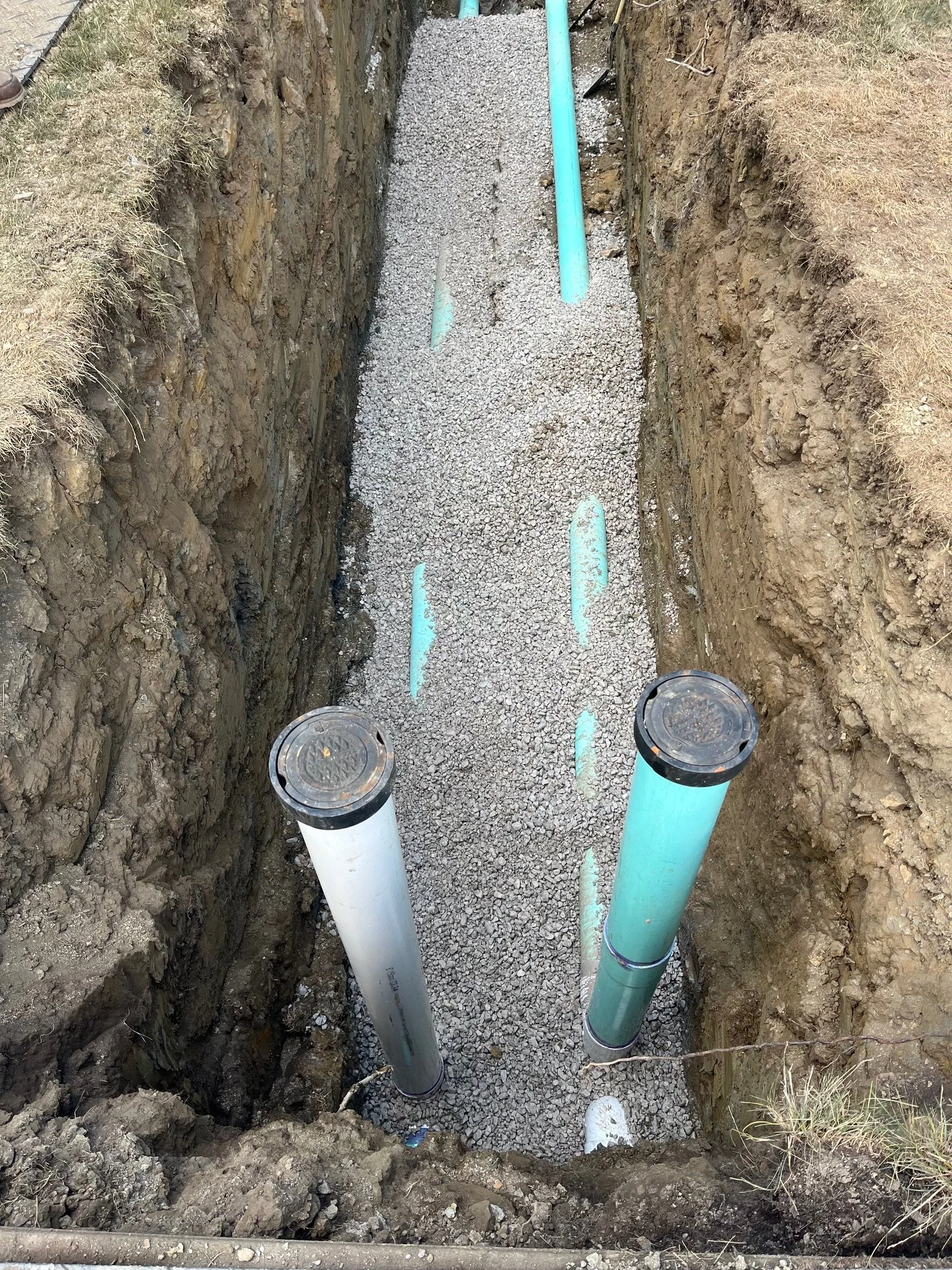 Underground utility installation with four aqua-colored pipes in a narrow trench with soil sides, gravel at the bottom, one white pipe, and one black cover on top of two pipes.