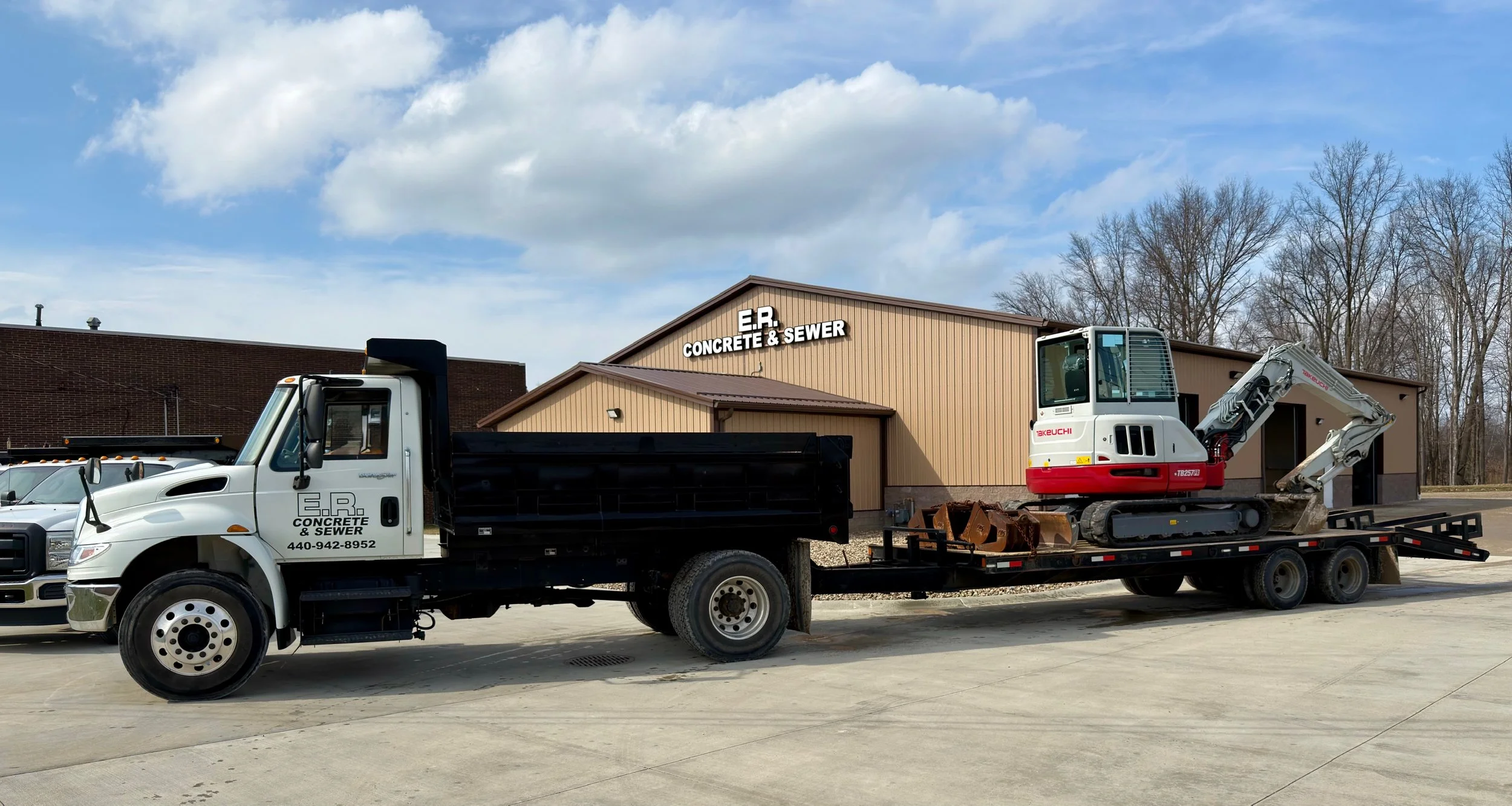 A flatbed truck parked outside a building labeled E.R. Concrete & Sewer, with a small excavator loaded on the flatbed.