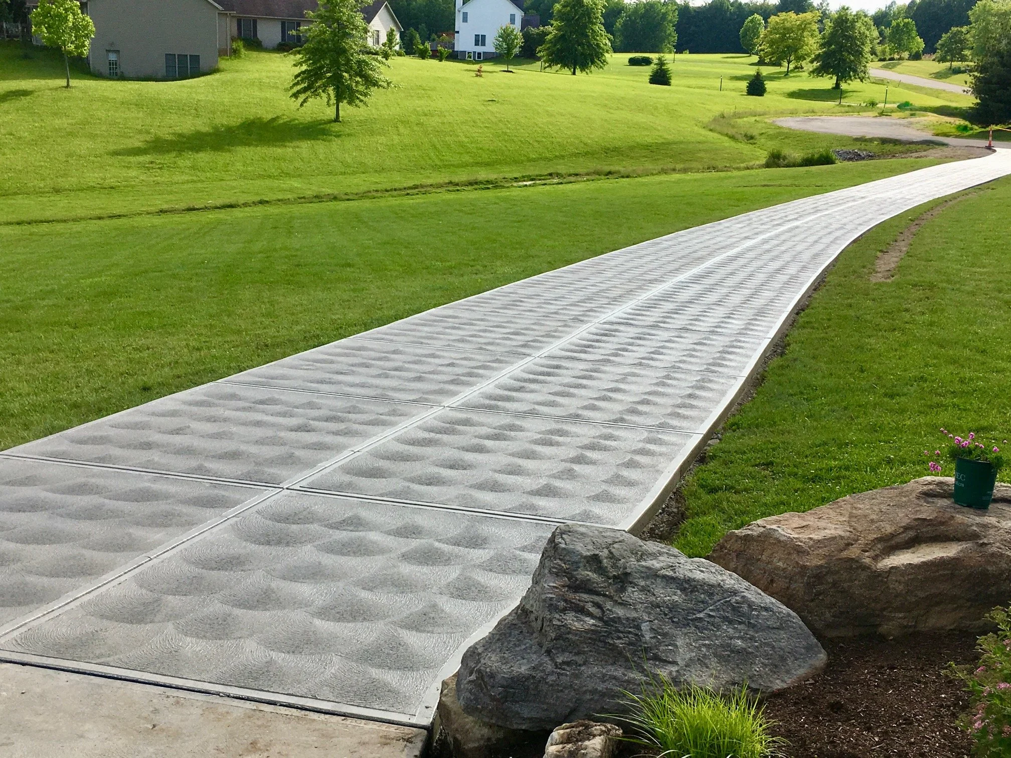 Concrete sidewalk with textured surface winding through a green yard with trees and houses in the background.
