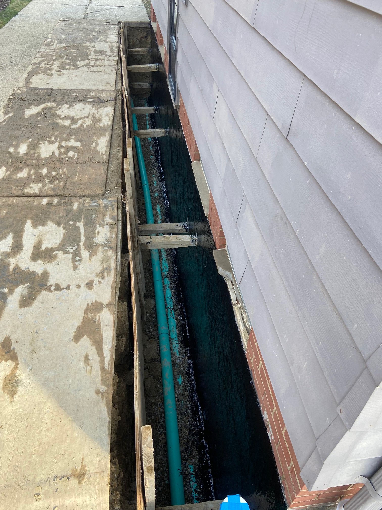 Construction waterproofing site showing a narrow trench next to a house with a gray siding exterior, a brick base, and an exposed blue plumbing pipe.