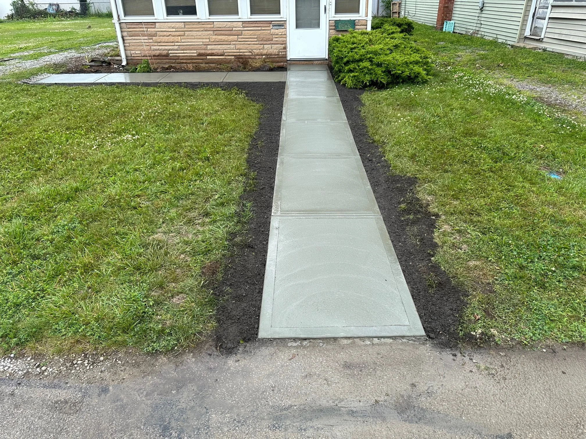 Freshly poured concrete sidewalk leading to the entrance of a house with brick and siding exterior, bordered by grass and soil.