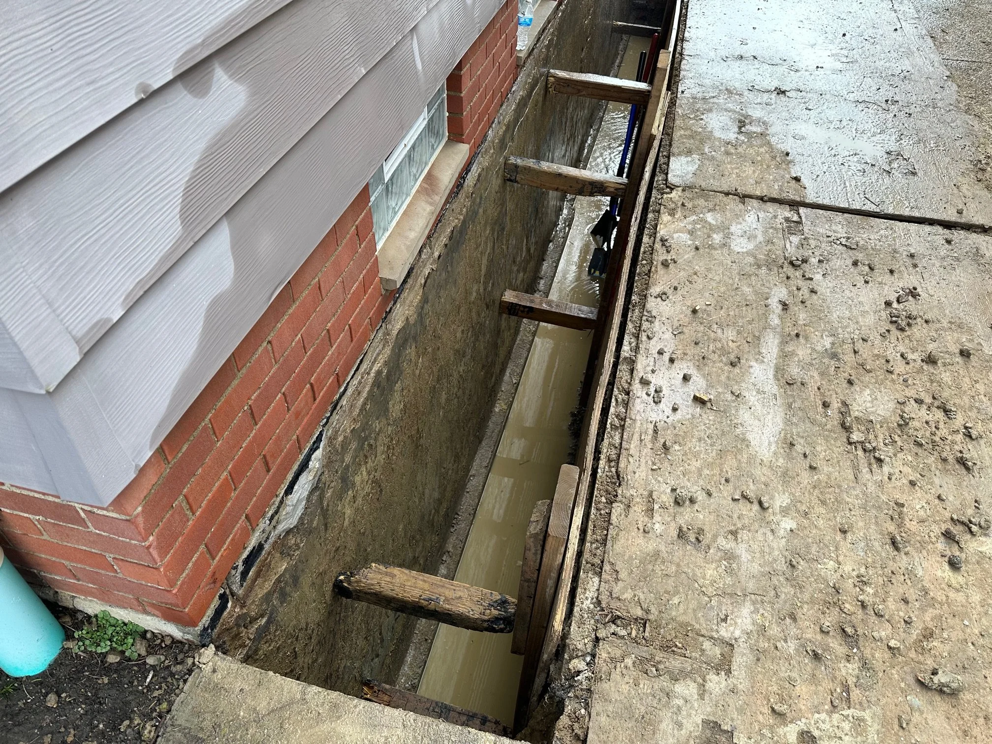 Construction waterproofing site along the side of a house, with a deep trench dug for a new foundation, supported by wooden posts and formwork, next to brick and siding exterior walls.