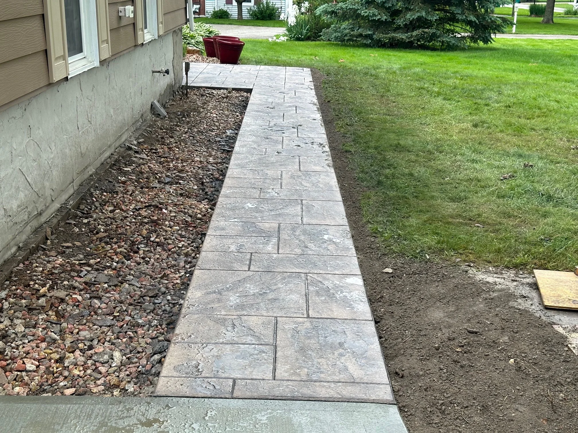 A newly installed concrete walkway next to a house, with a bed of small rocks on one side and a green lawn on the other, in a residential yard.