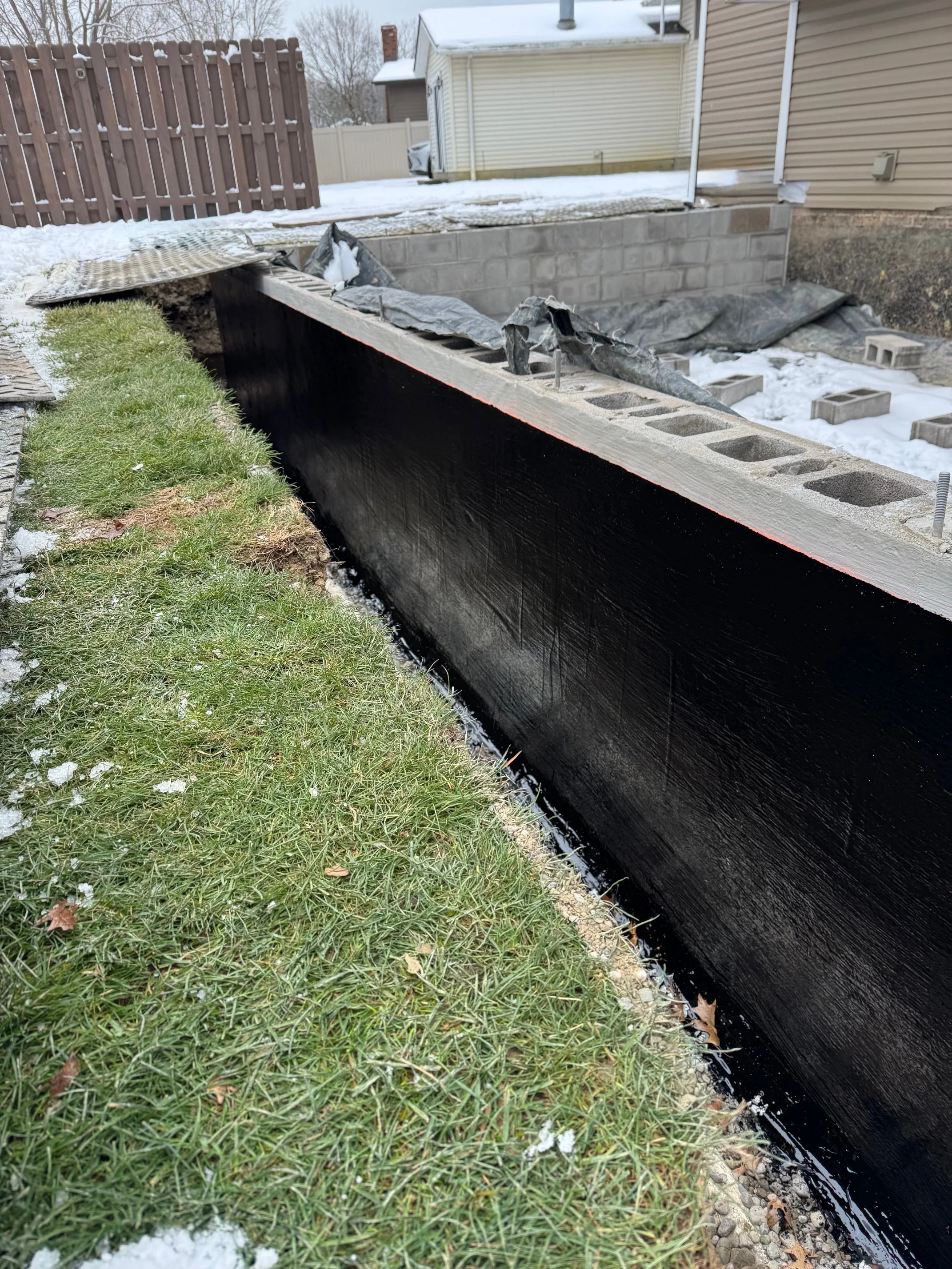 Construction site with cinder blocks, black waterproofing on the foundation, and green grass with some snow in the foreground.