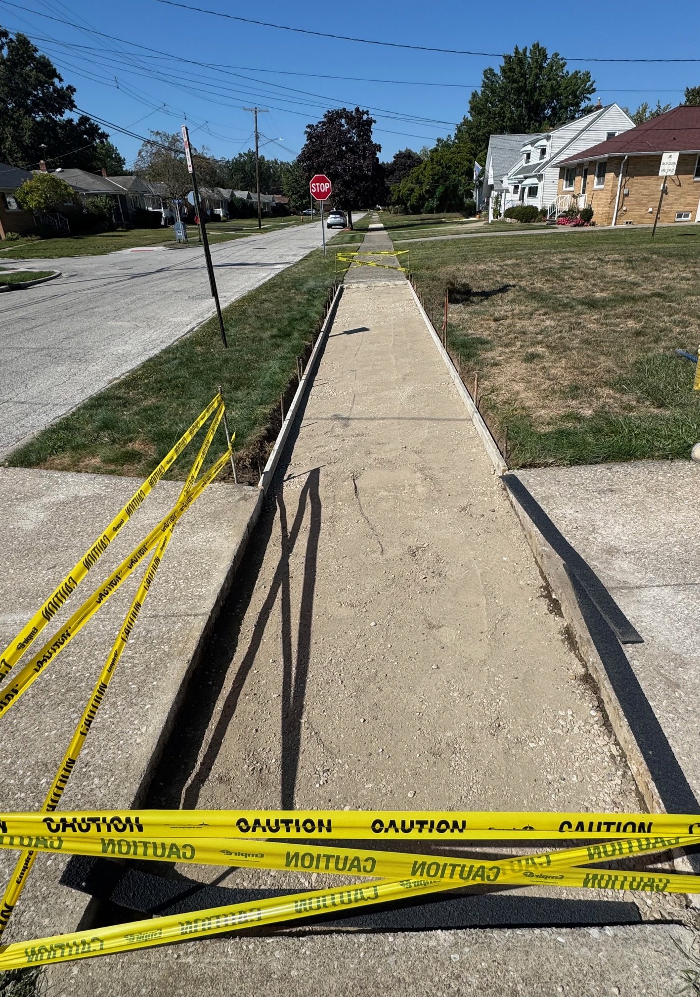 Sidewalk under construction with yellow caution tape, a stop sign in the distance, residential houses, green grass, and a clear blue sky.