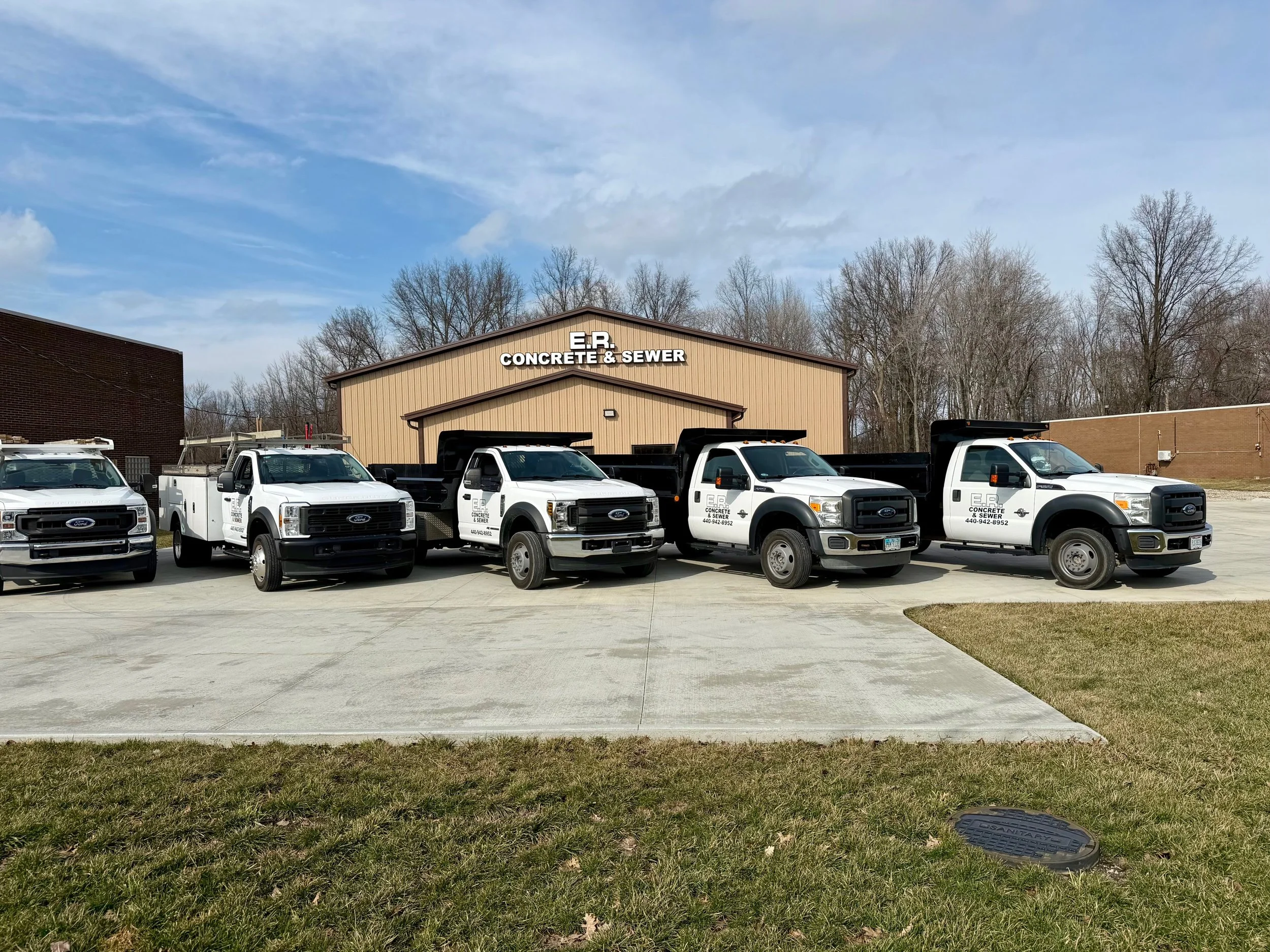 Four white Ford trucks with black dump beds parked in front of a building labeled 'E.R. Concrete & Sewer'.