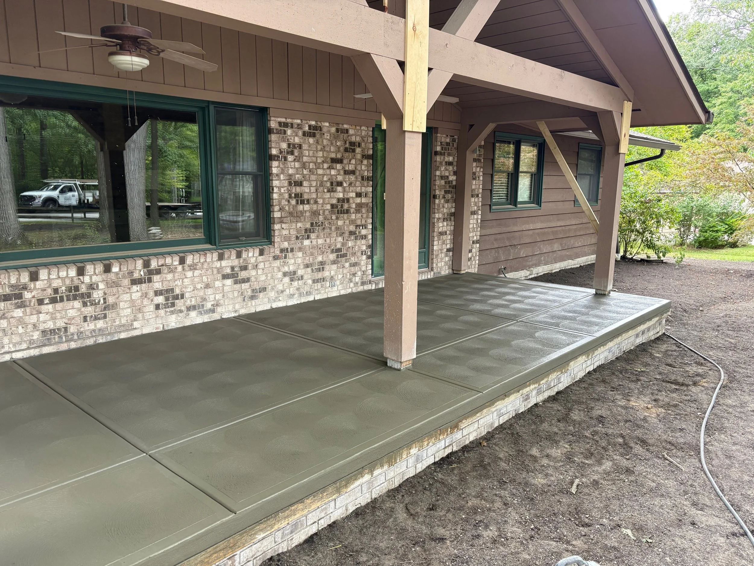 Newly poured concrete patio with textured surface outside the back of a house with brick and wood exterior, under a covered porch with ceiling fan and windows.