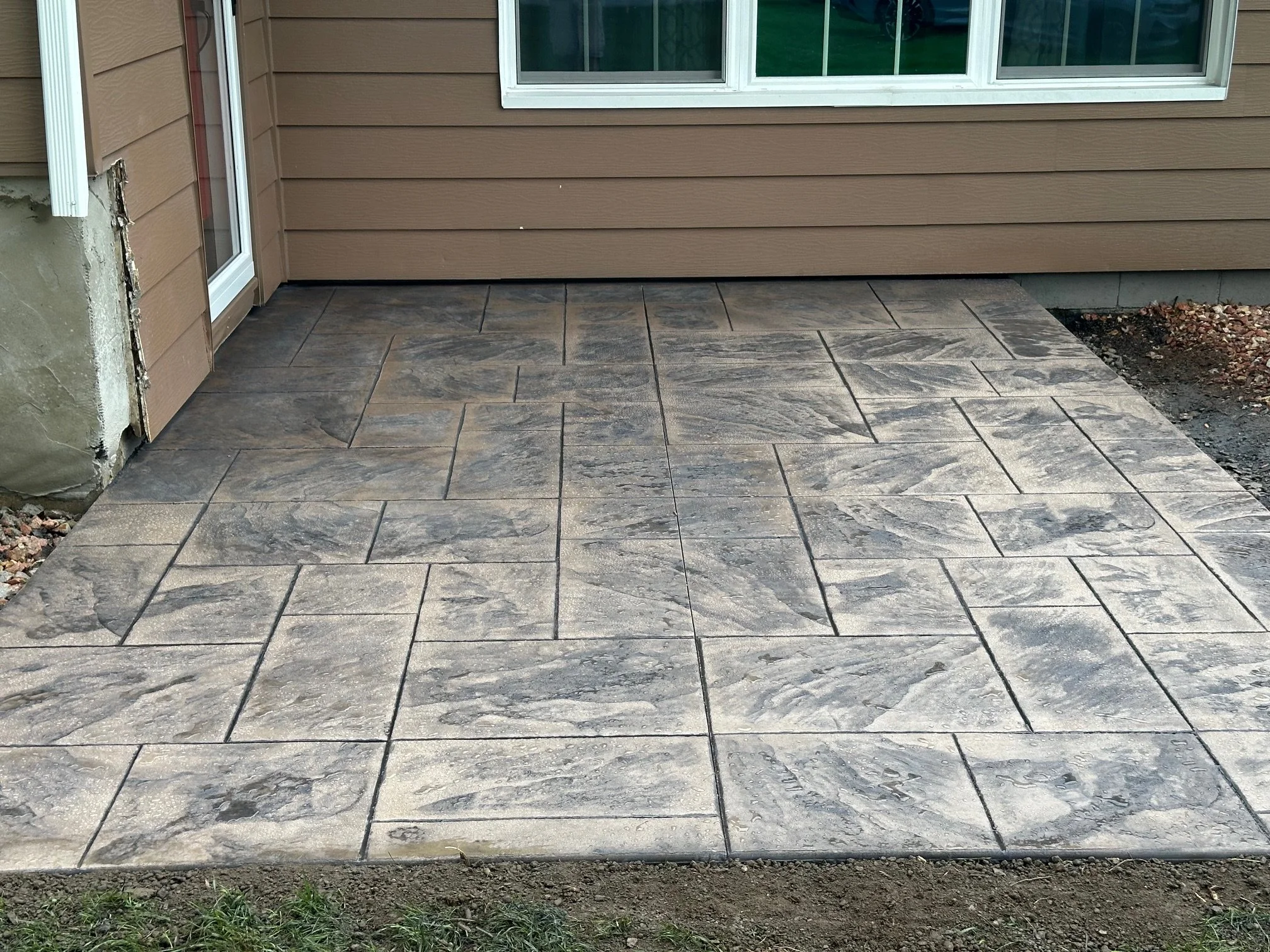 Newly installed stone patio with rectangular stone tiles in front of a house with brown siding, a window, and a sliding glass door.