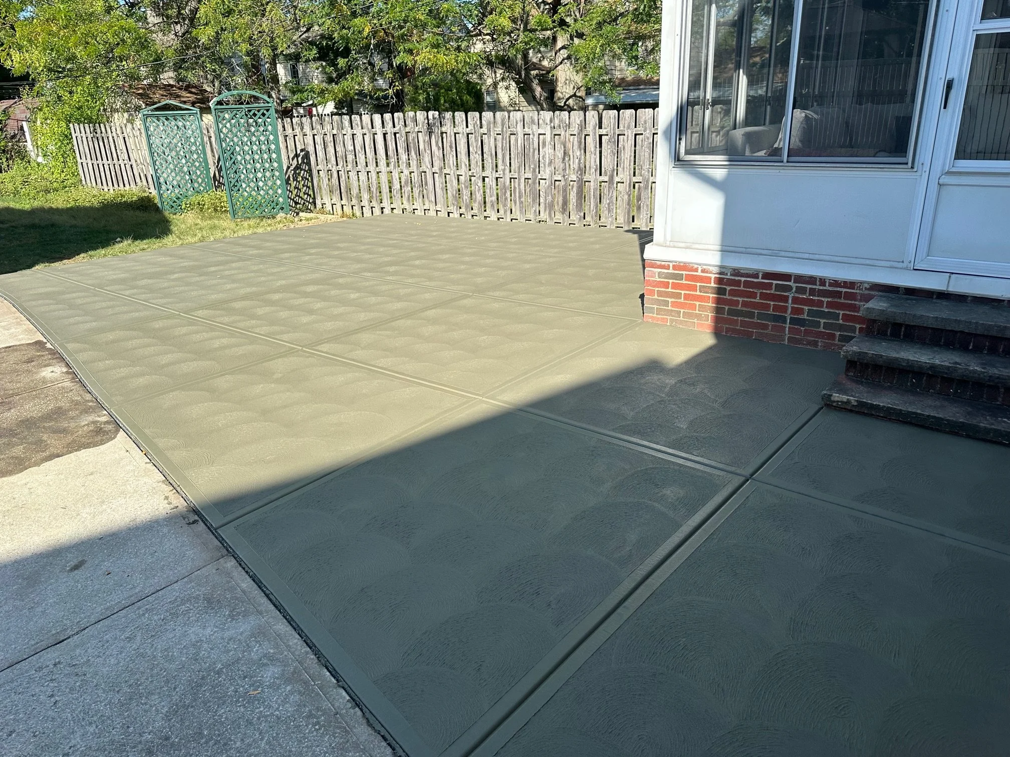 Newly poured concrete patio with expansion joints next to a house and a wooden fence, in a backyard.