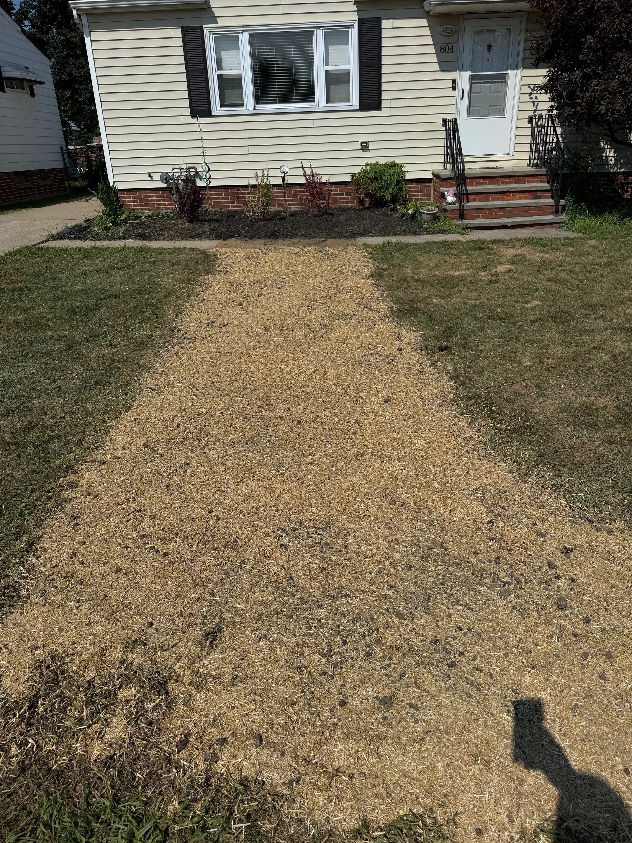 Front yard with a gravel pathway leading to a house with a white door, a window with black shutters, and a small garden bed with plants and decorative items.