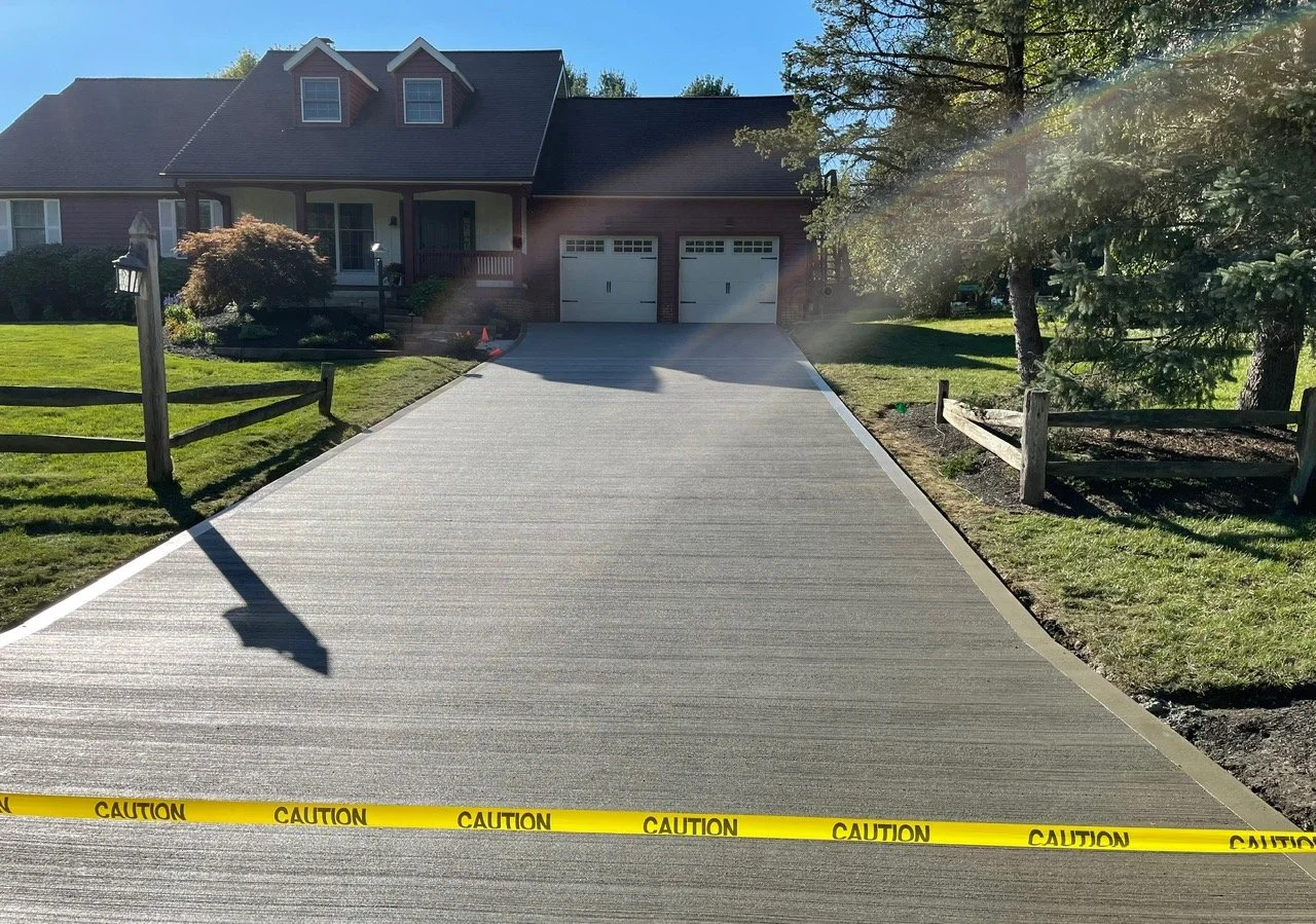 Freshly poured concrete driveway leading to a house with a two-car garage, bordered by a lawn and trees, with caution tape at the foreground.