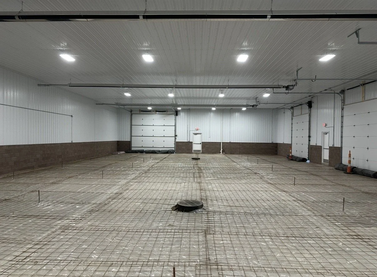 Interior of a large building under construction, with exposed rebar on the floor, metal walls, ceiling with multiple lights, and a garage door at the back.