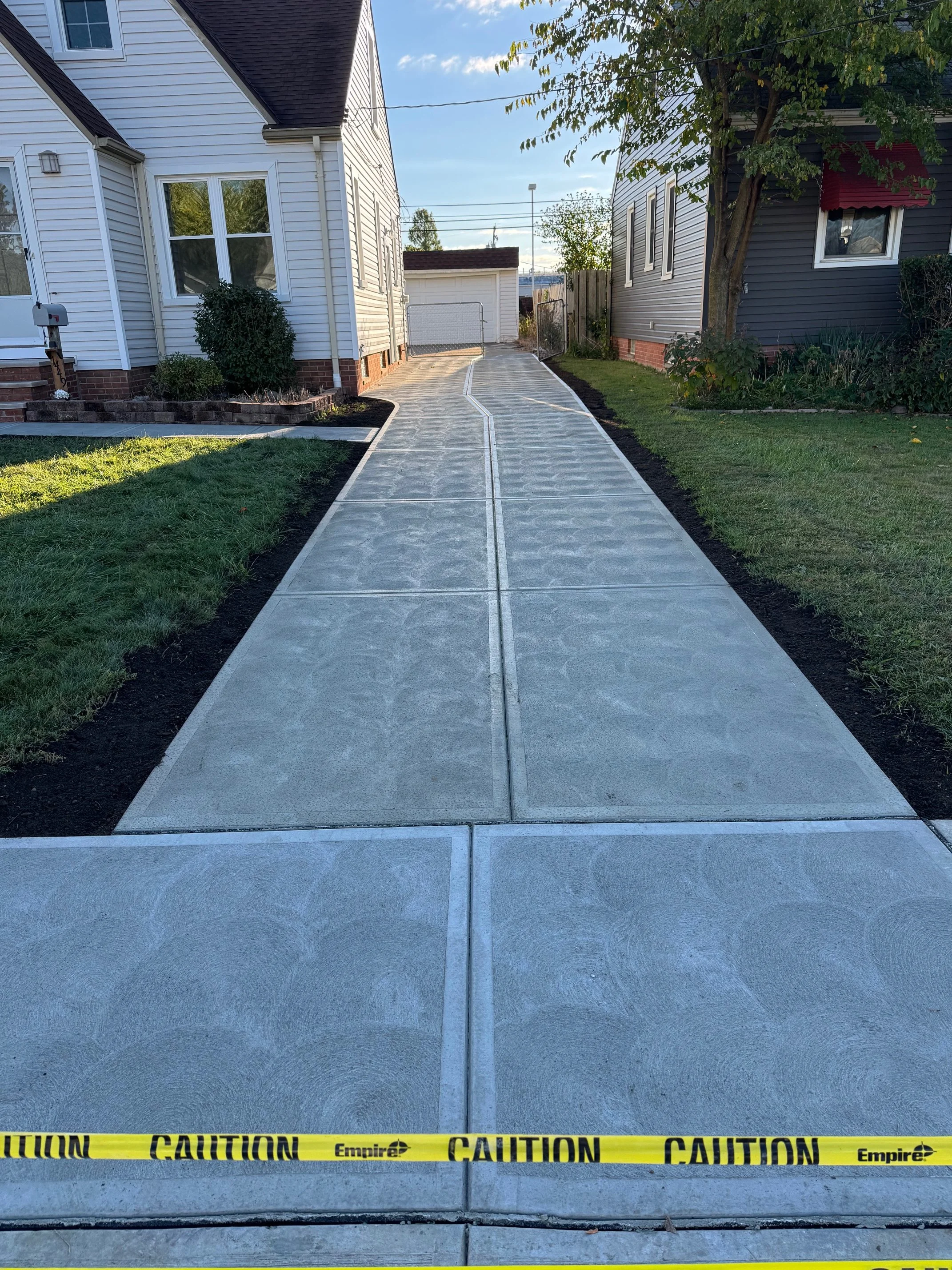 Concrete driveway with newly poured sections, bordered by grass and soil, with caution tape across the front. Houses and a garage are visible in the background on a sunny day.