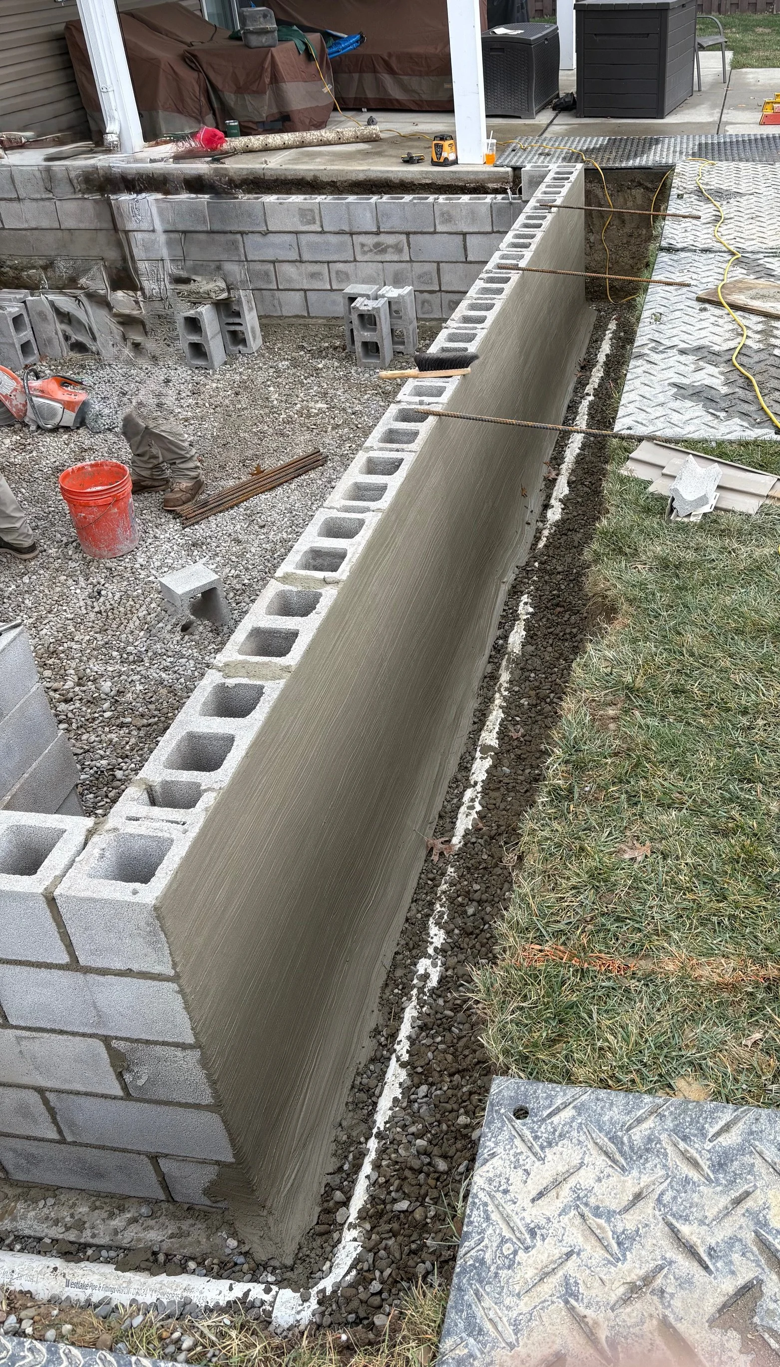 Construction site with a partially built cinder block wall and freshly applied cement, outdoor setting with tools, construction materials, and gravel ground.