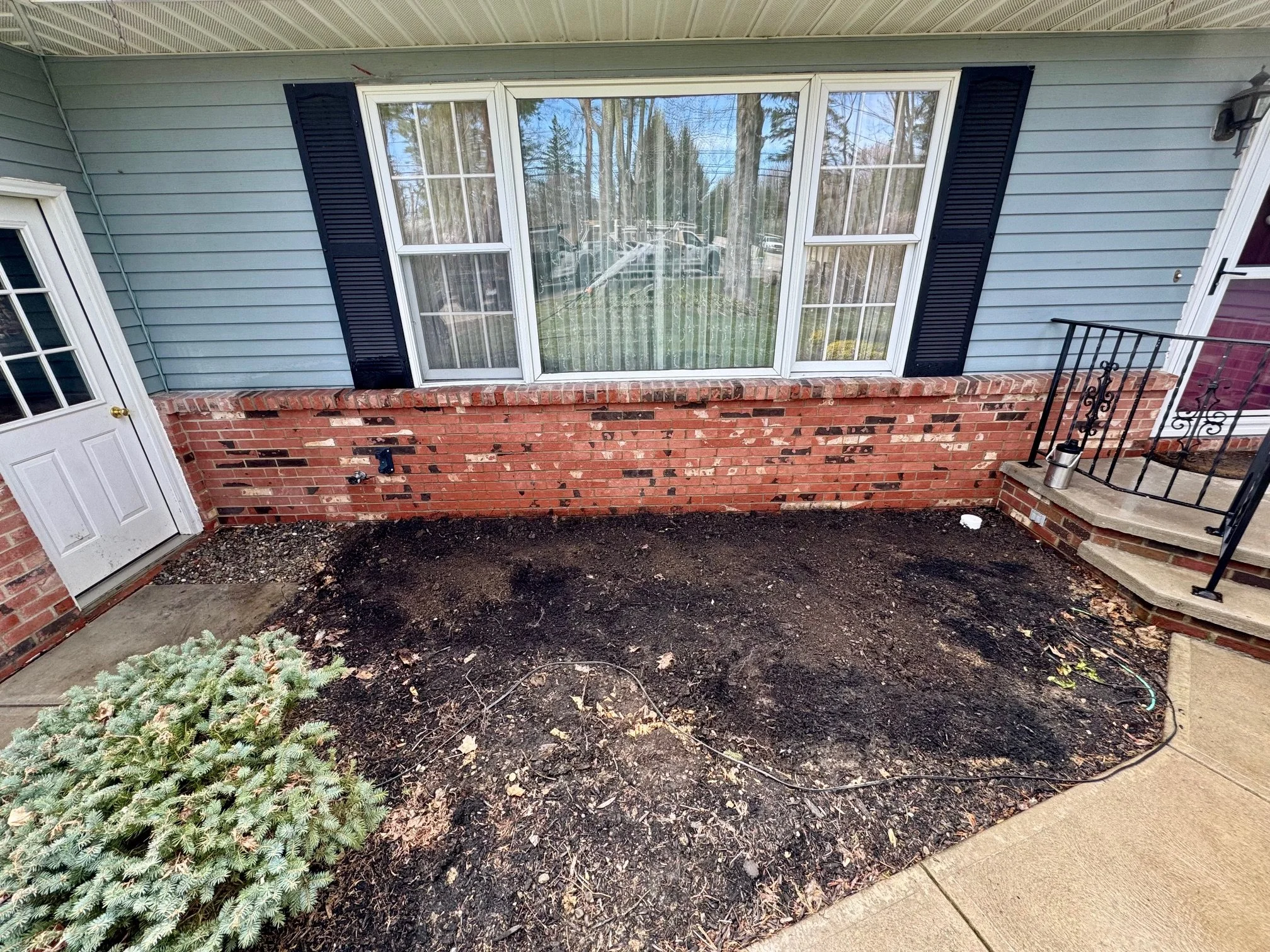 Exterior porch area with dirt and mulch where plants may be planted, a small bush in the corner, a brick wall, a large window, a white door with glass panels, black shutters, a small set of steps with a black railing, and a porch light.