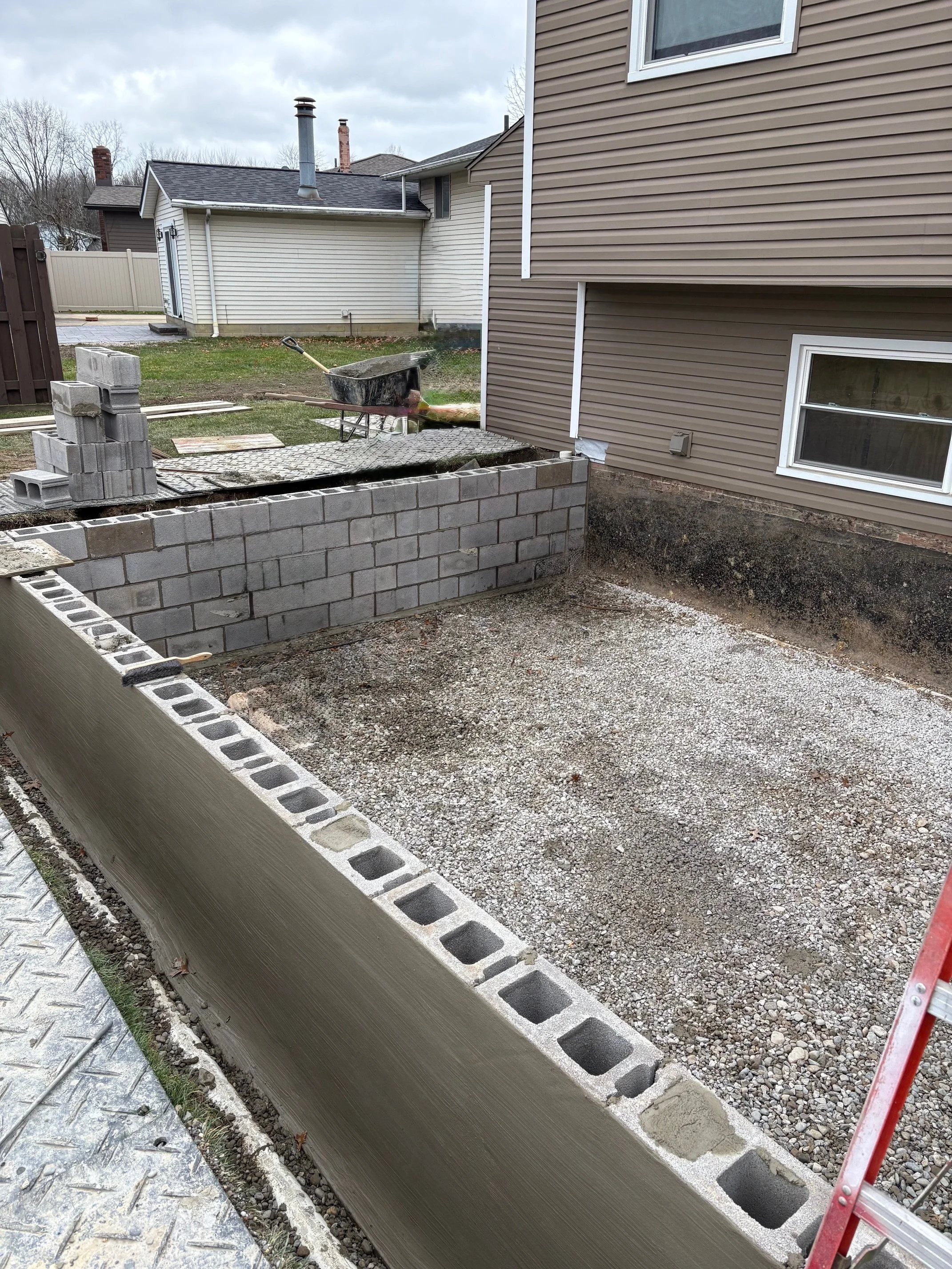 Construction site with cinder blocks forming a building foundation, gravel ground, a wheelbarrow, and fitting tools near residential houses.