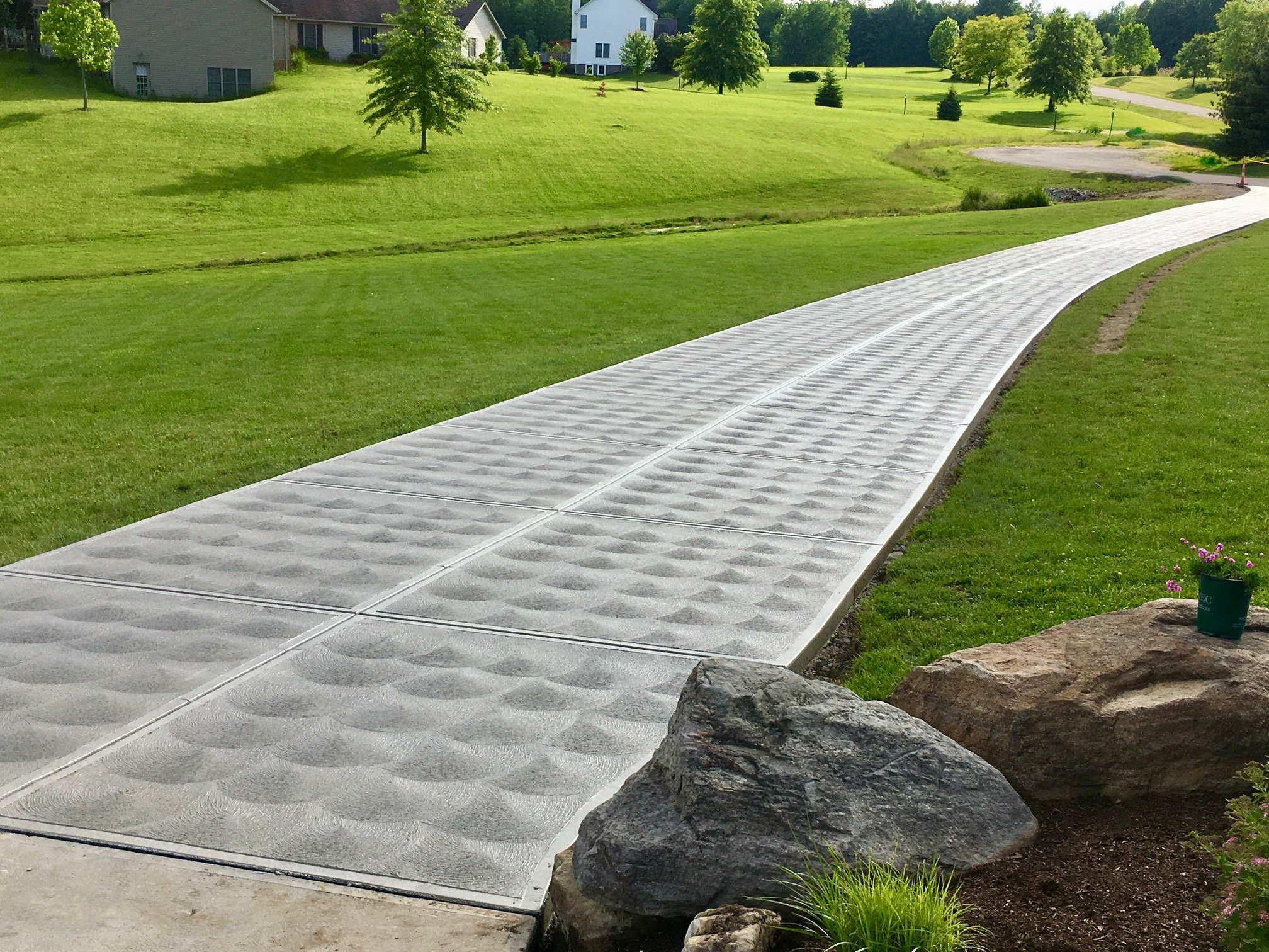 Newly installed concrete sidewalk with textured surface in a suburban neighborhood, surrounded by a well-maintained lawn and trees.