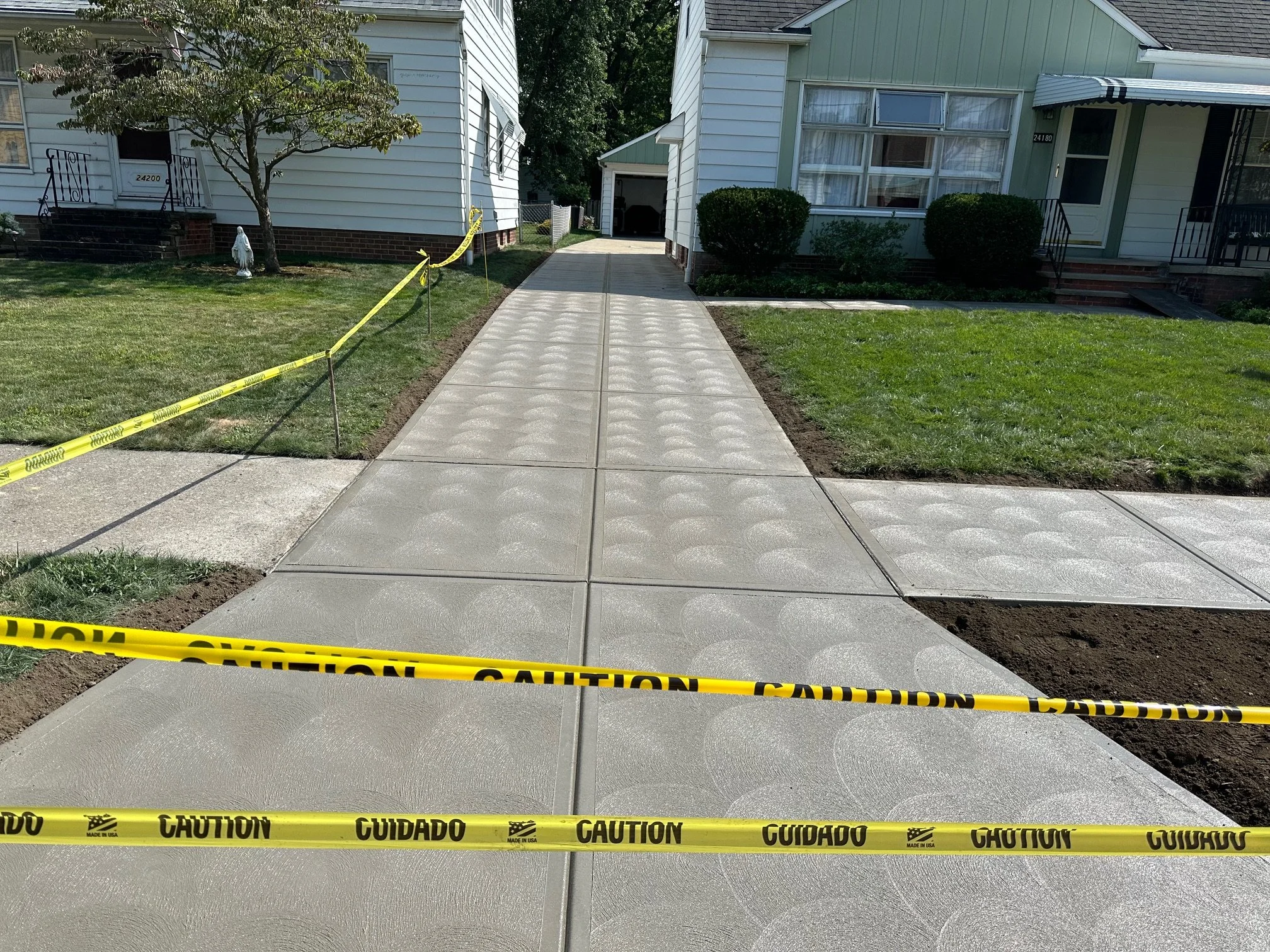 Newly poured concrete sidewalk section in front of a house, with caution tape surrounding the area, indicating ongoing construction or installation.