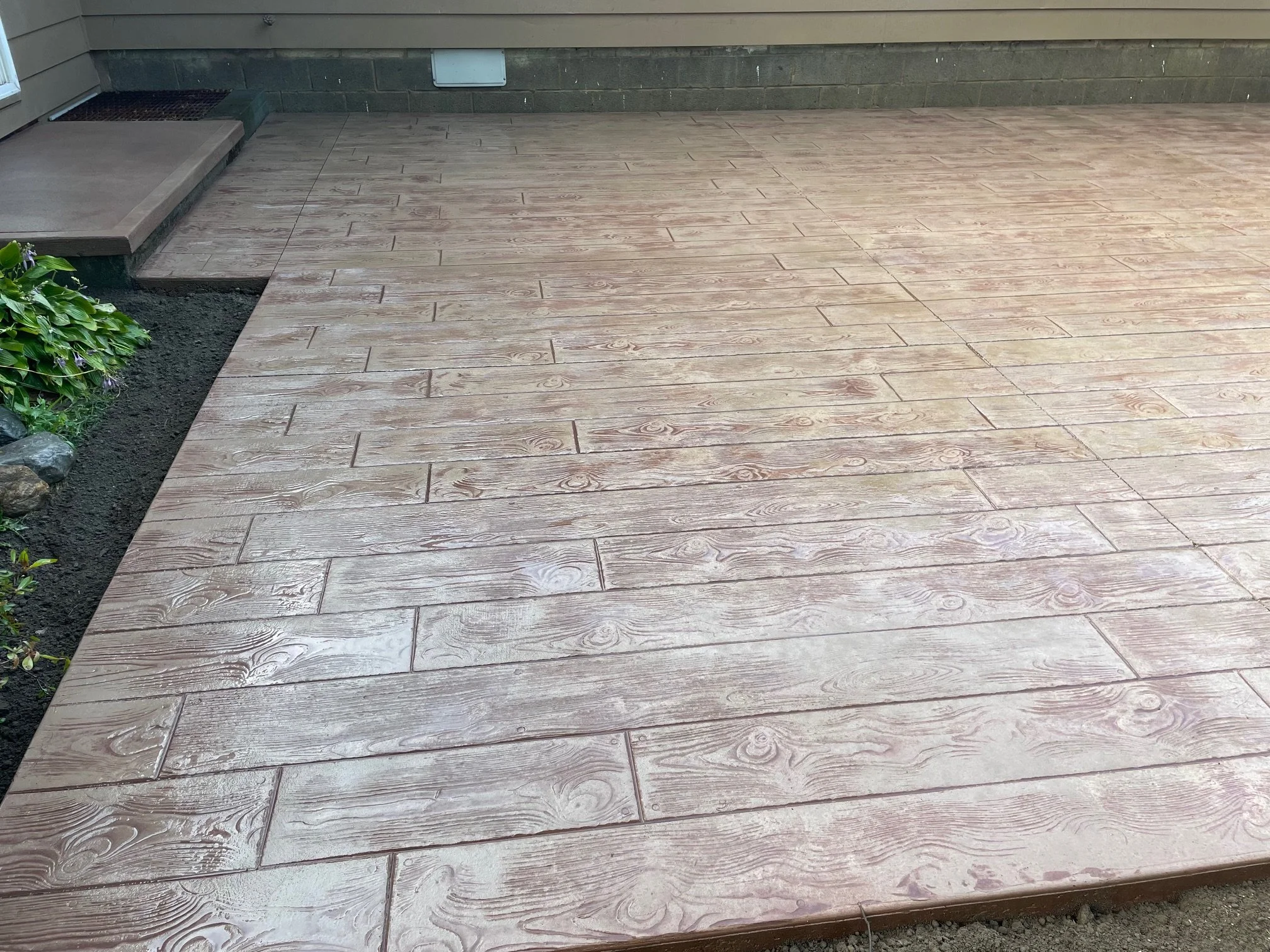 Wood-look outdoor patio with new wooden planks, adjacent to flowerbed and small steps leading to a door, home exterior visible.