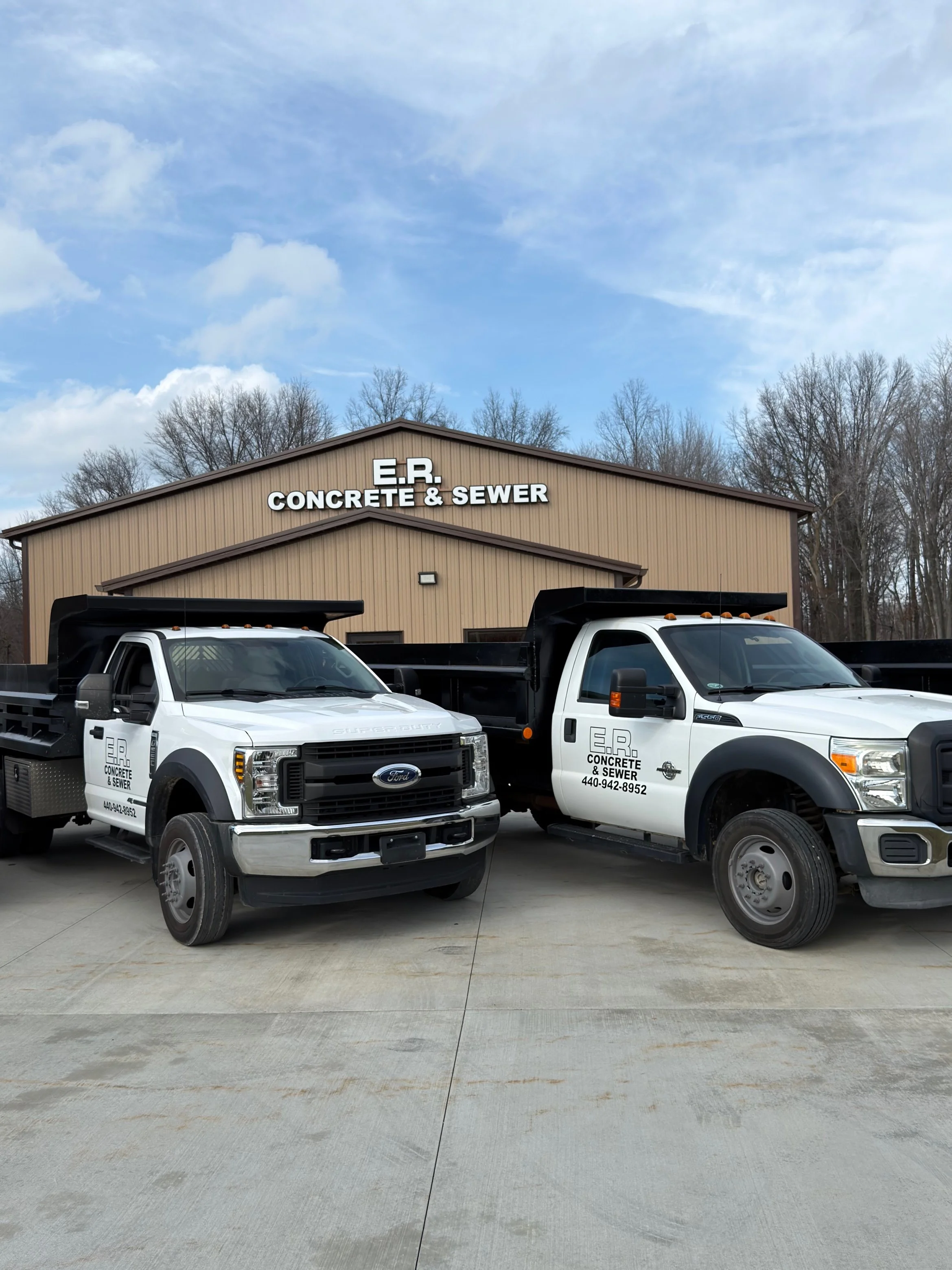 Two white trucks parked in front of a beige building with a sign that reads "E.R. CONCRETE & SEWER." The trucks have company logos and contact information on the doors.