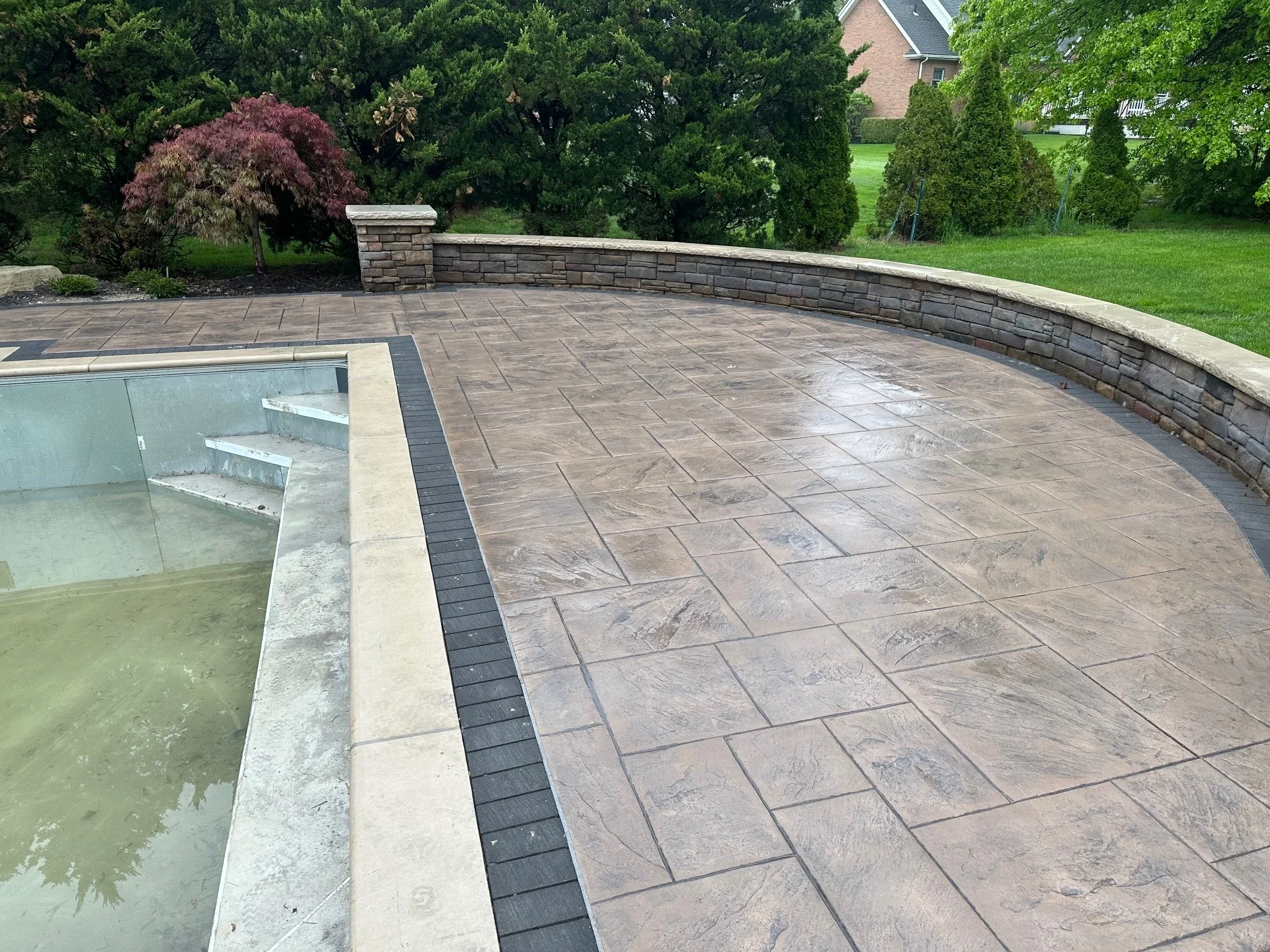 Backyard patio with stone tiles, a stone retaining wall, and a swimming pool with steps, surrounded by green trees and grass.