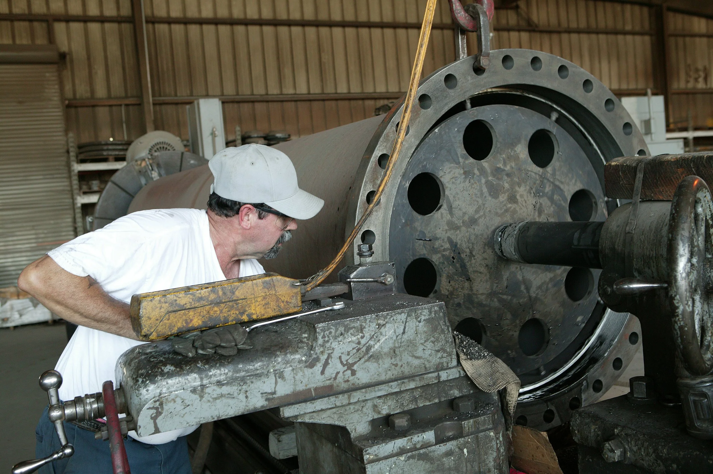 A man wearing a white cap and glasses working on a large industrial metal component inside a workshop.