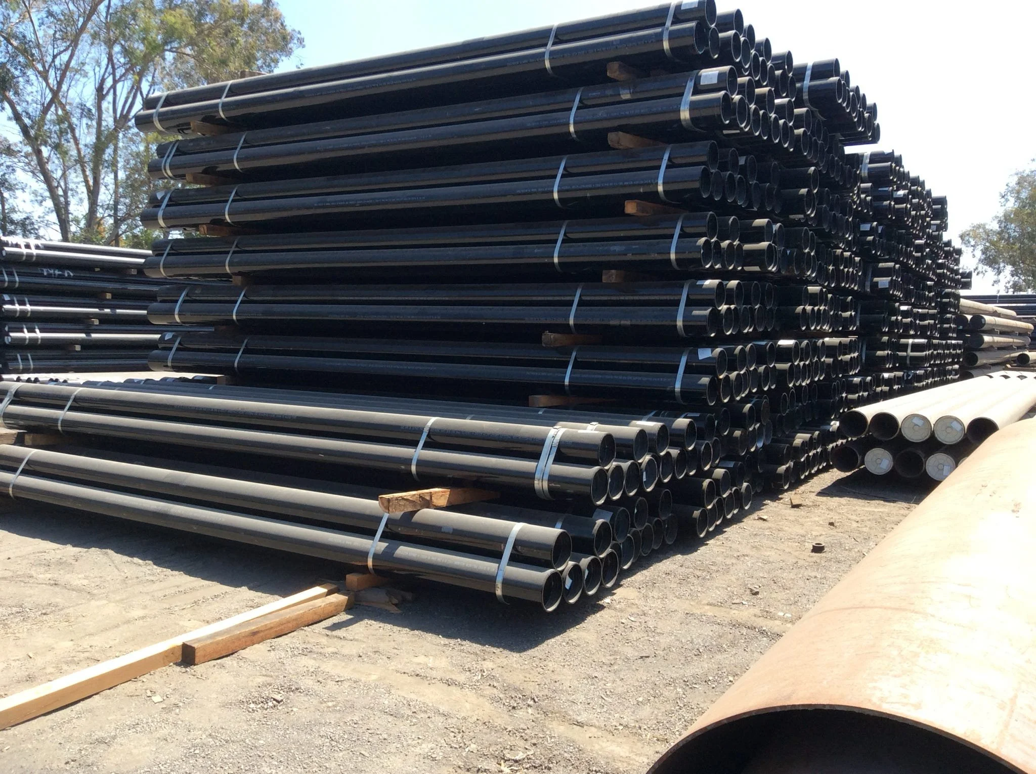 Stacked black steel pipes secured with straps on wooden pallets outdoors.
