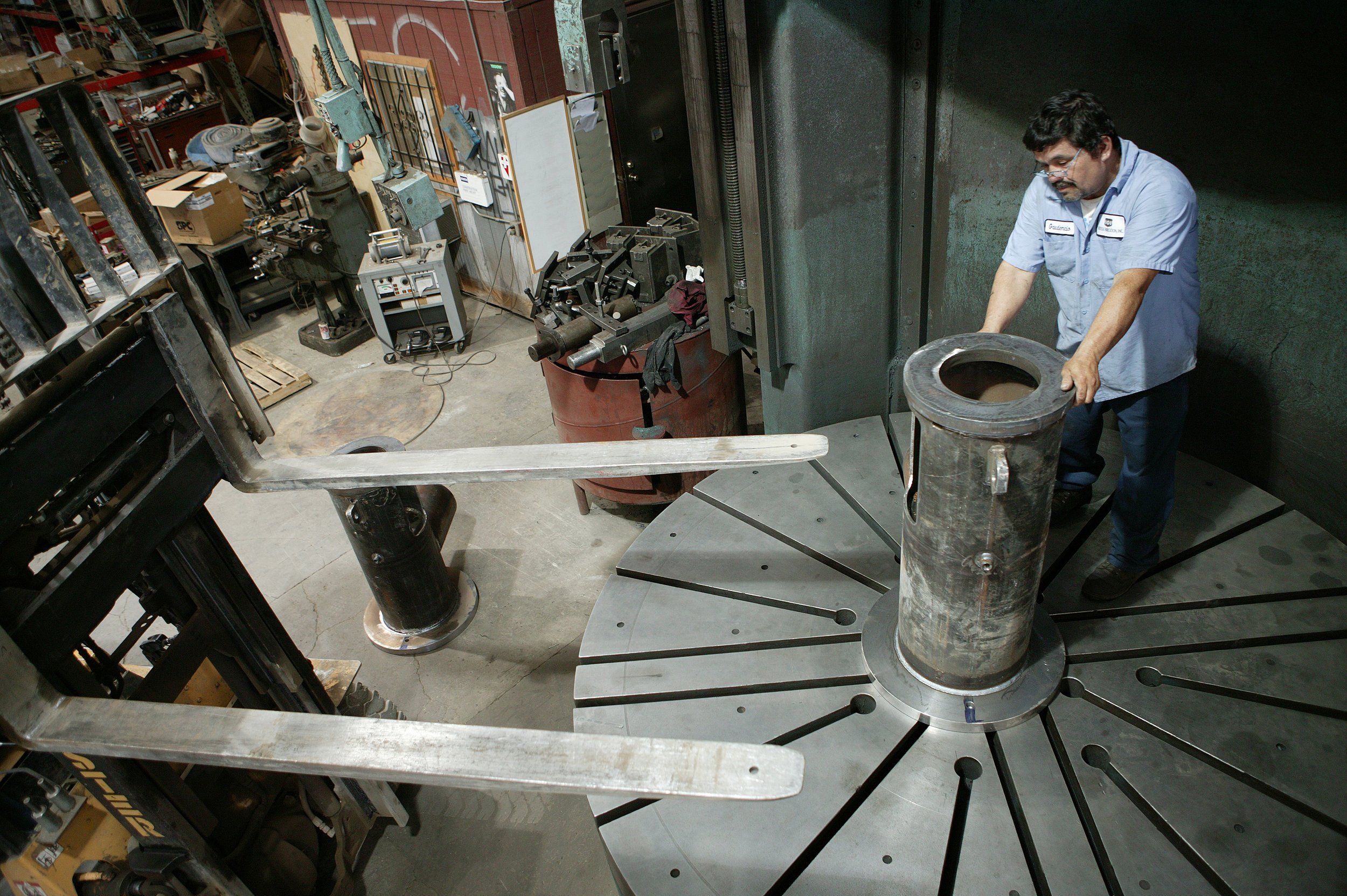 A man wearing glasses and a light blue work shirt is working in a workshop, handling a metal ring or pipe over a metal table. The workshop has various tools, equipment, and boxes on shelves and workbenches in the background.
