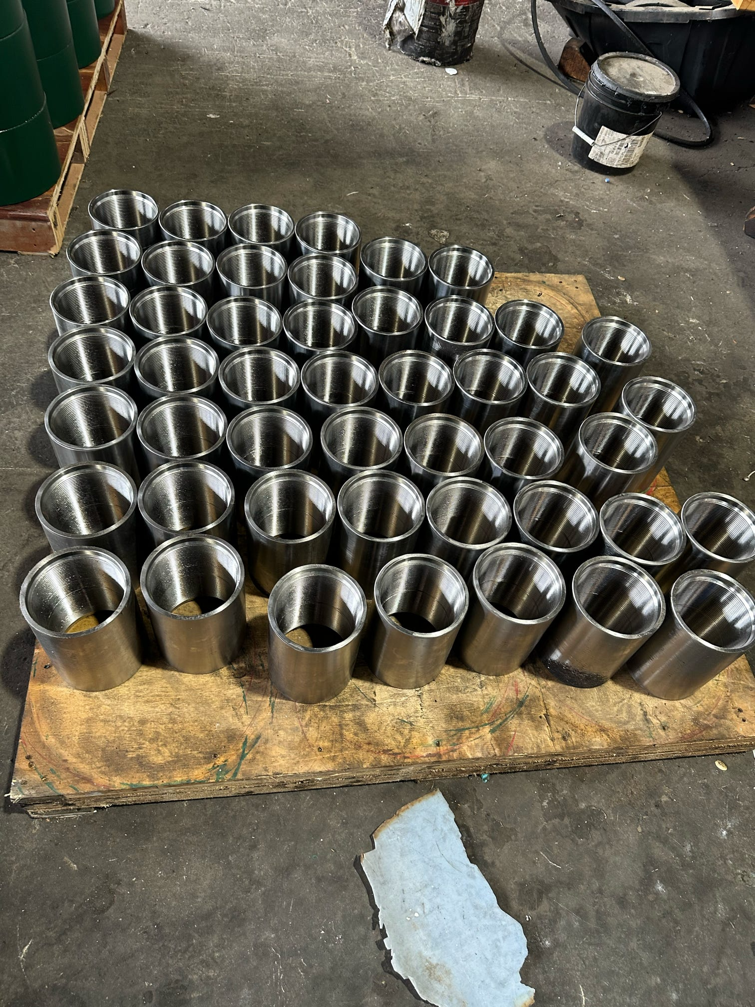 A workshop scene with a wooden workbench holding multiple shiny metal cylinders arranged in rows. The background shows a black container, a bucket, and a work surface with various tools and materials. The concrete floor has a painted white line and s