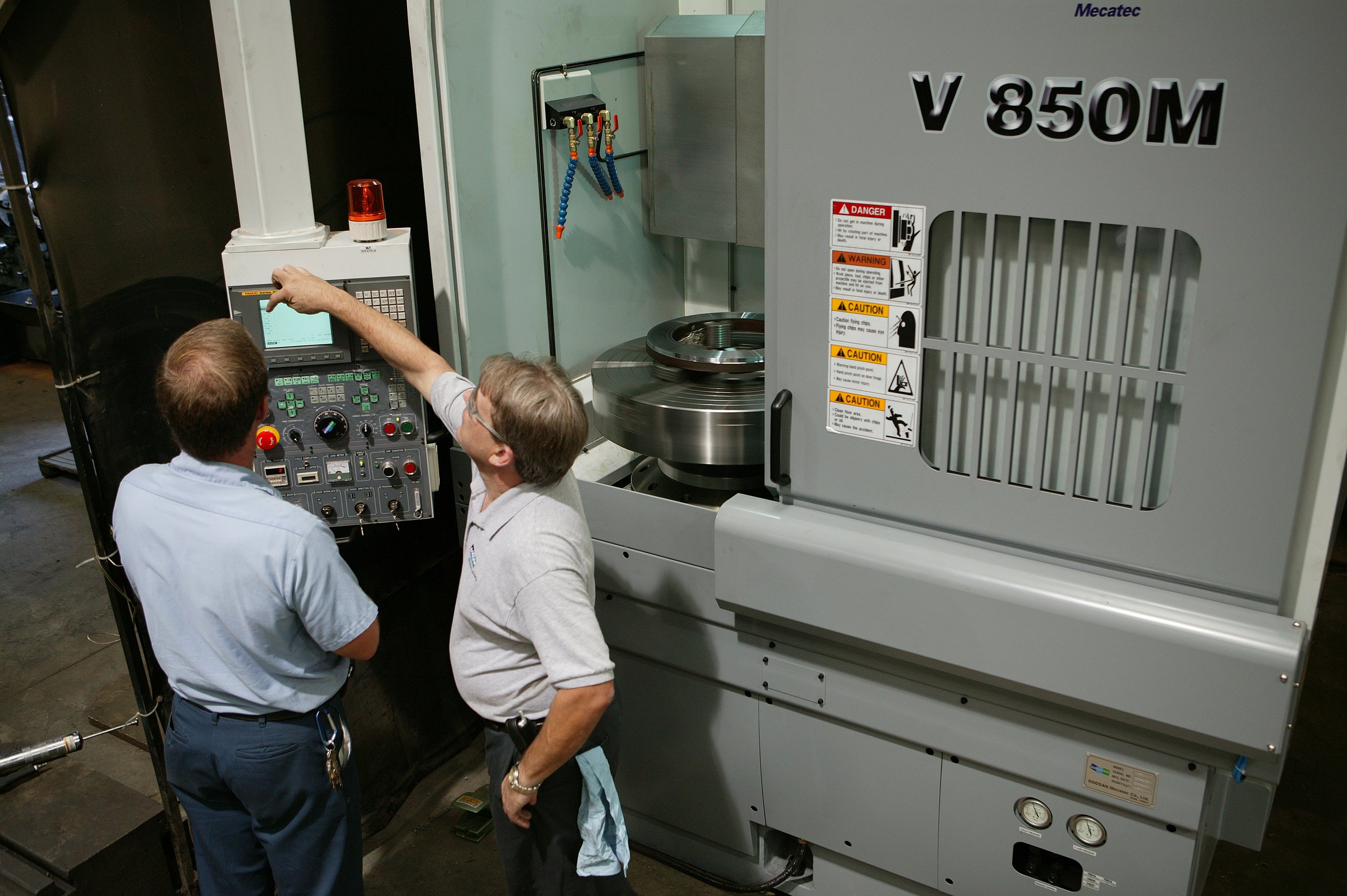 Two men operating large industrial machinery labeled V 850 M in a factory setting.