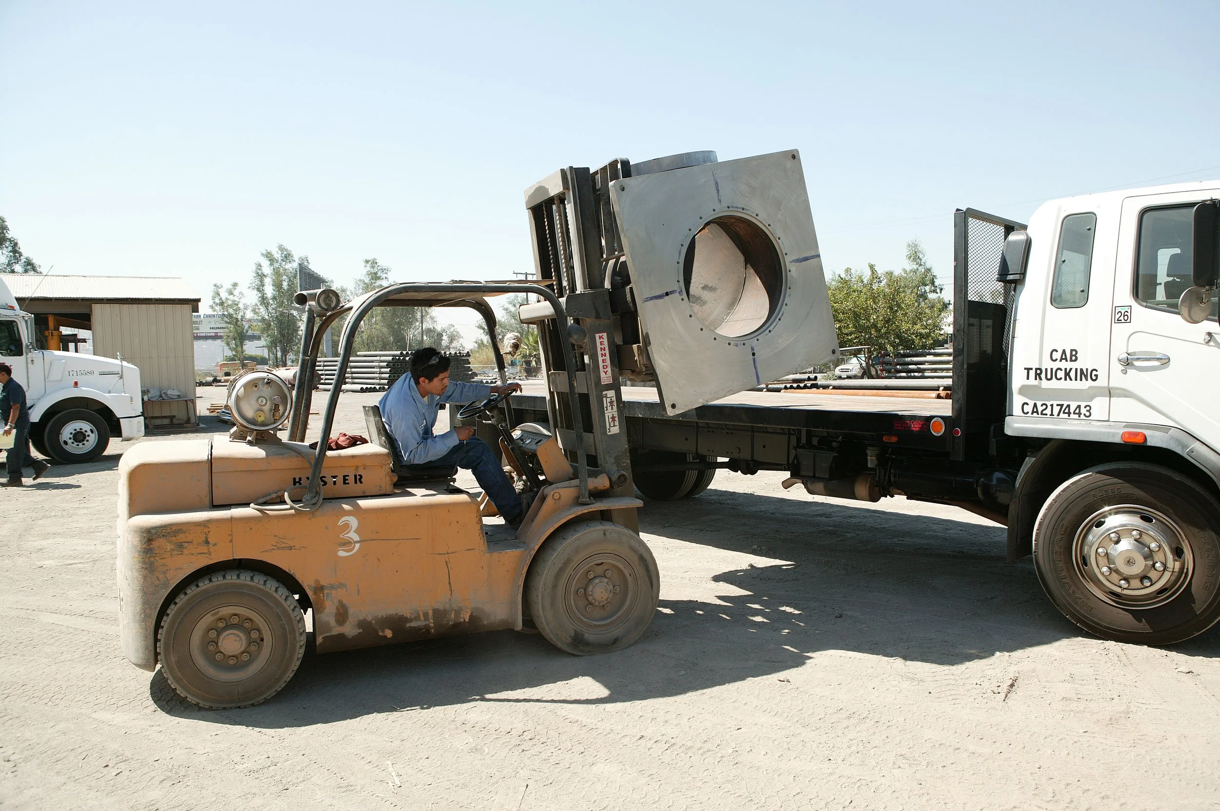 A worker operating a forklift with a large metal component on a flatbed truck, across an outdoor industrial yard.