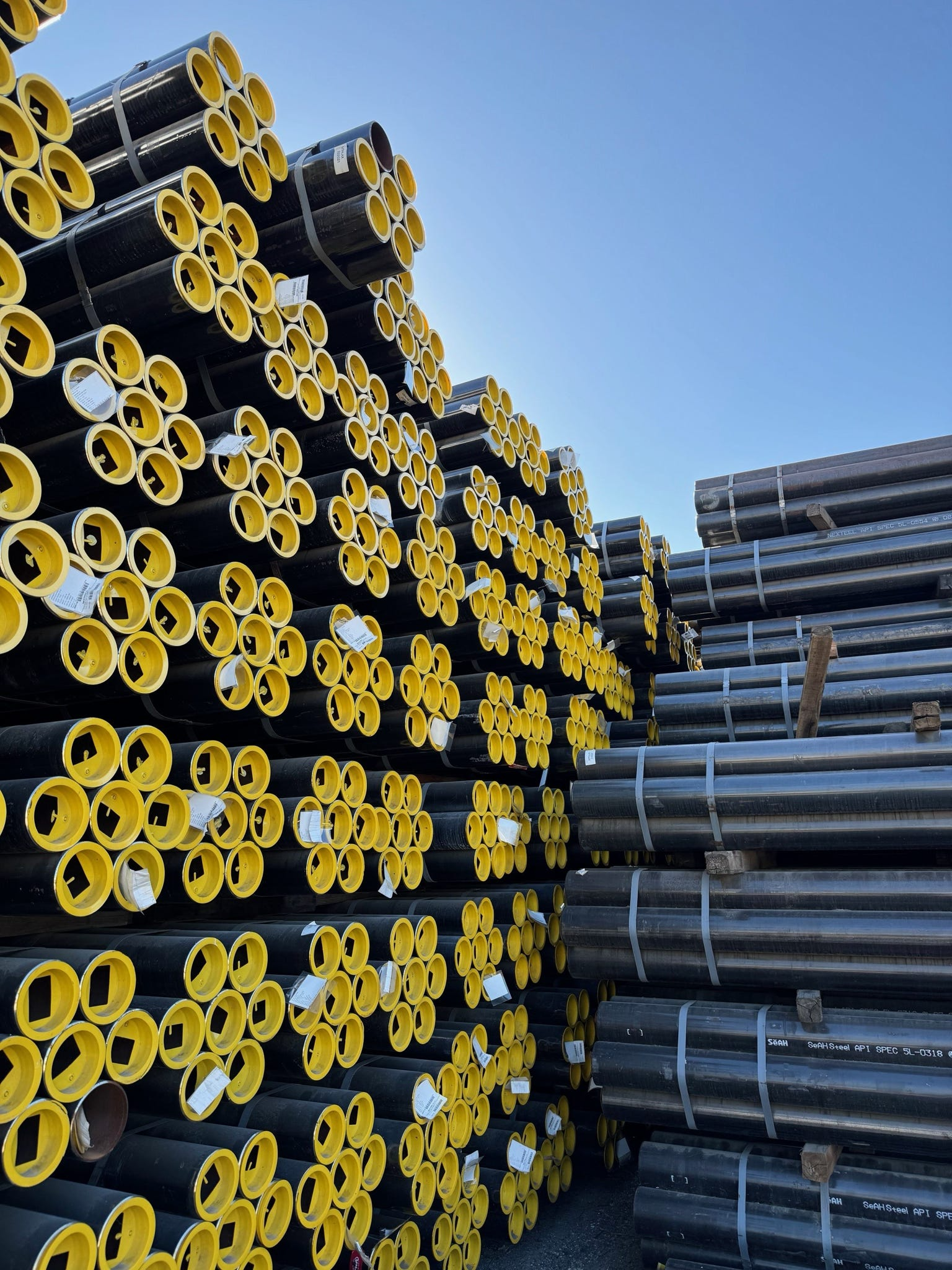 Stacks of black and yellow steel pipes, bundled and organized on pallets outdoors under a clear blue sky.