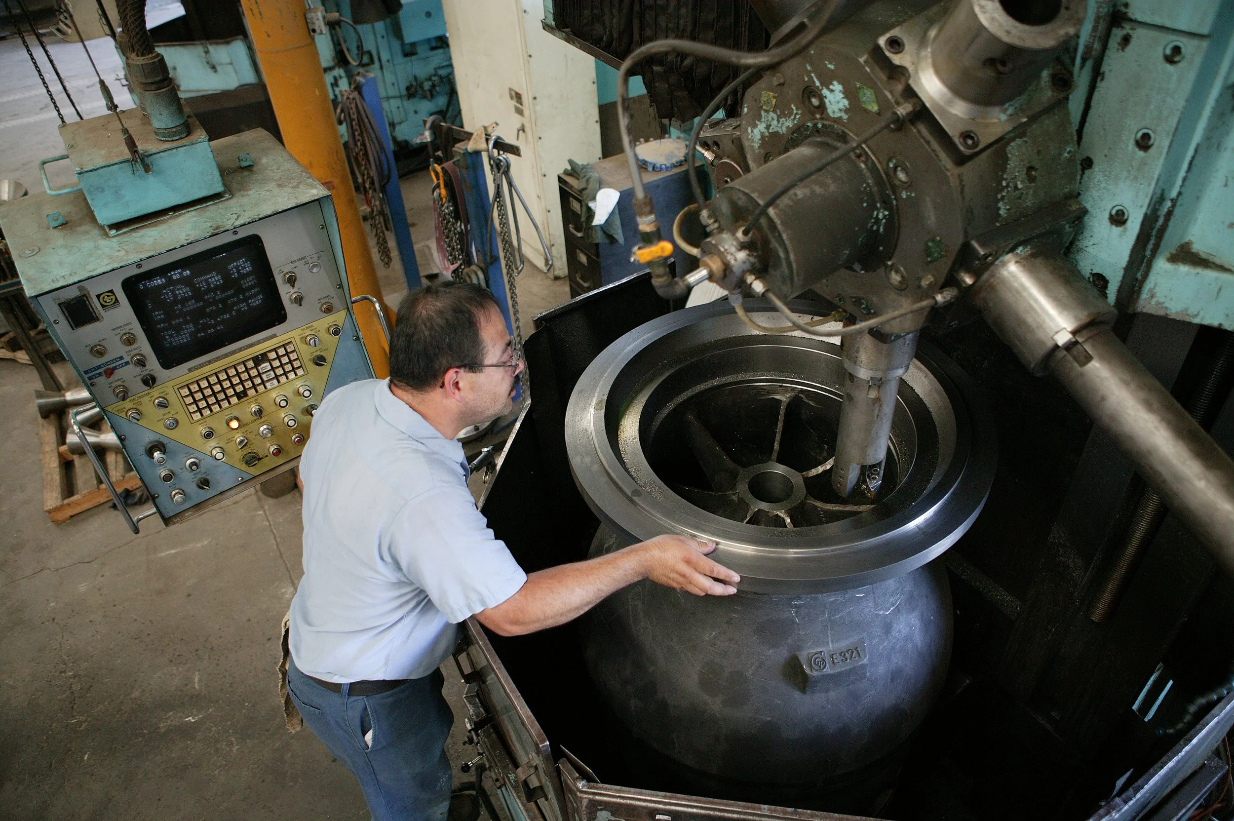 A man working with a large industrial machine, possibly a lathe or a CNC machine, in a workshop. The machine has a circular component, and the man is inspecting or adjusting something within it.