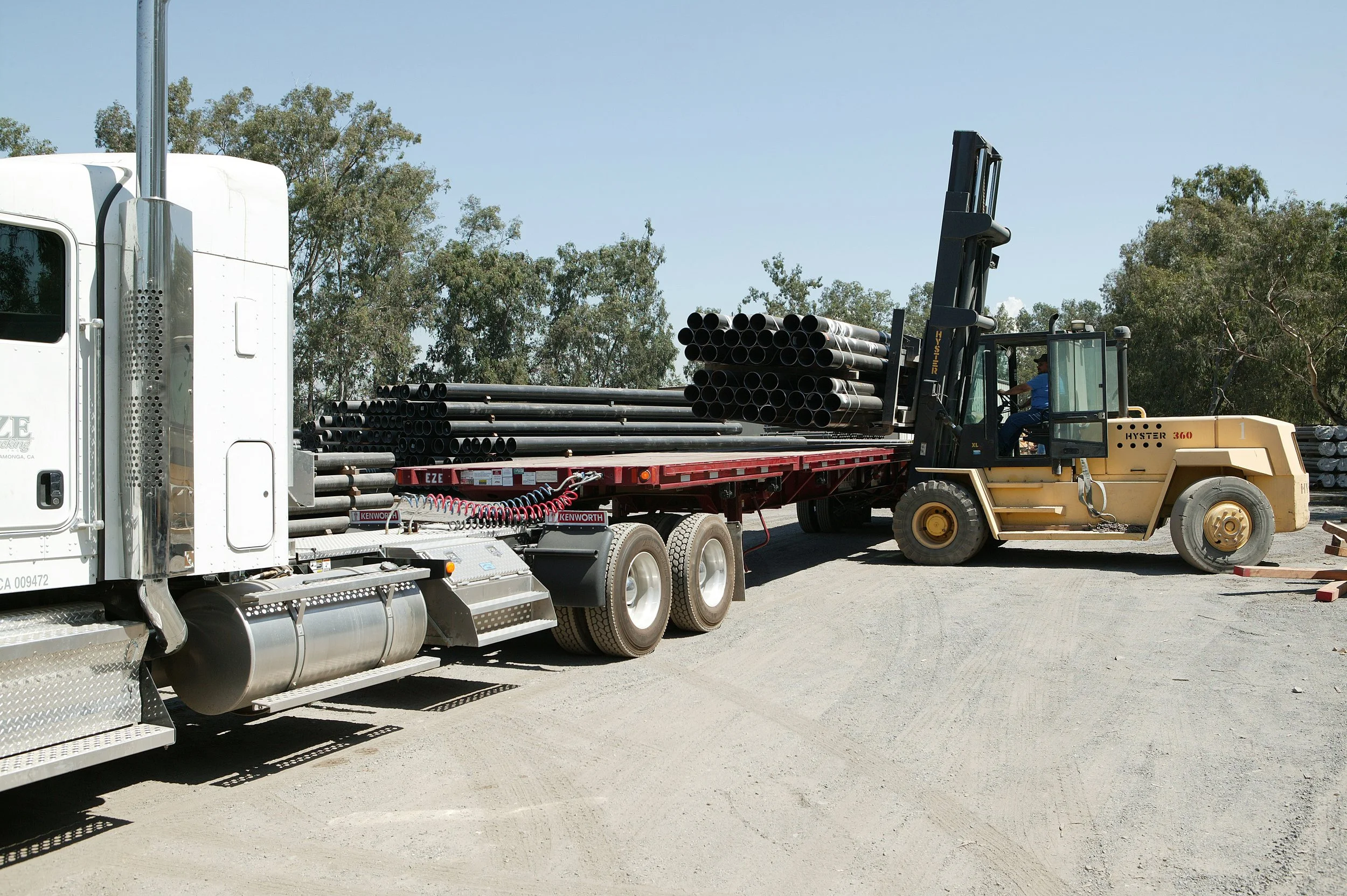 A large semi-truck with black pipes loaded on a flatbed trailer, with a forklift operator in a blue shirt moving the pipes. Trees and clear sky are in the background.