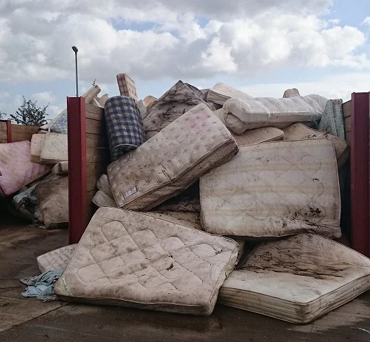 A pile of discarded old mattresses and box springs outside on a concrete surface, with a cloudy sky in the background.