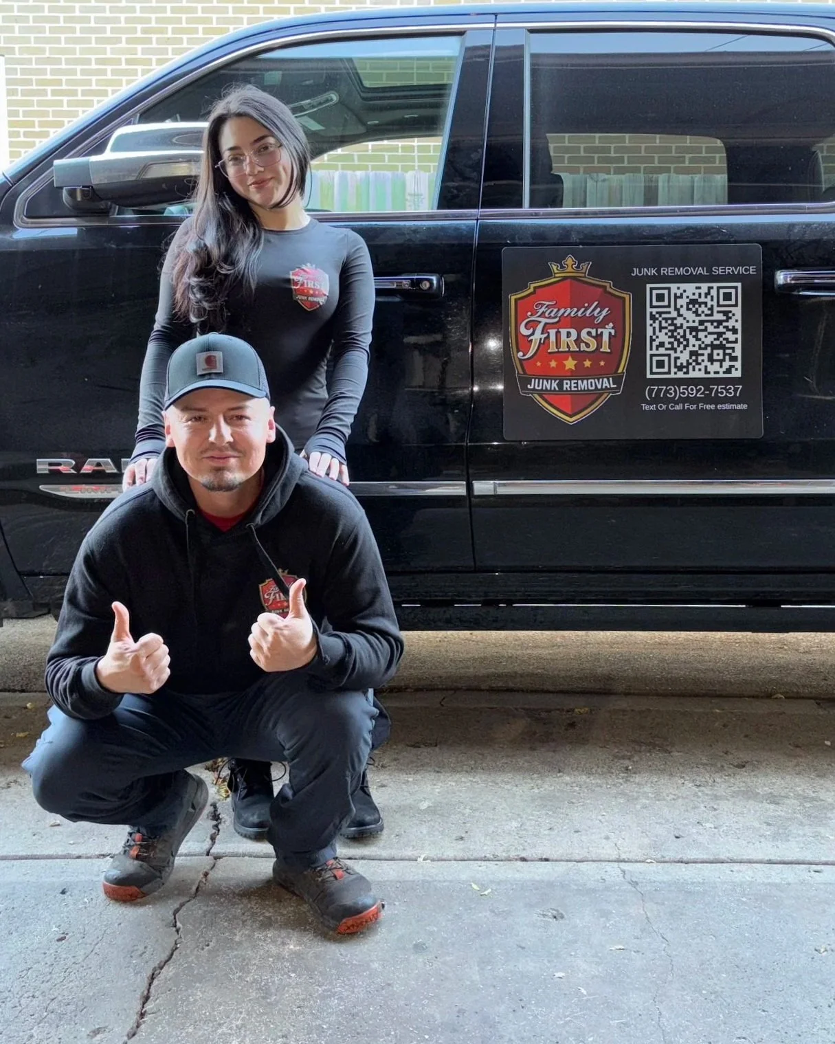 Two people, a woman and a man, in front of a black vehicle with a family first junk removal sign on the side. The man is crouching, giving two thumbs up, and wearing a black hoodie and cap. The woman is standing behind him, smiling, with her hands on his shoulders. Both are wearing branded clothing with the Family First logo.