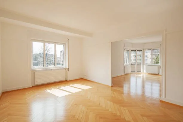 Empty living room with large windows, hardwood flooring, and a sunlit bay window area in an apartment or house.