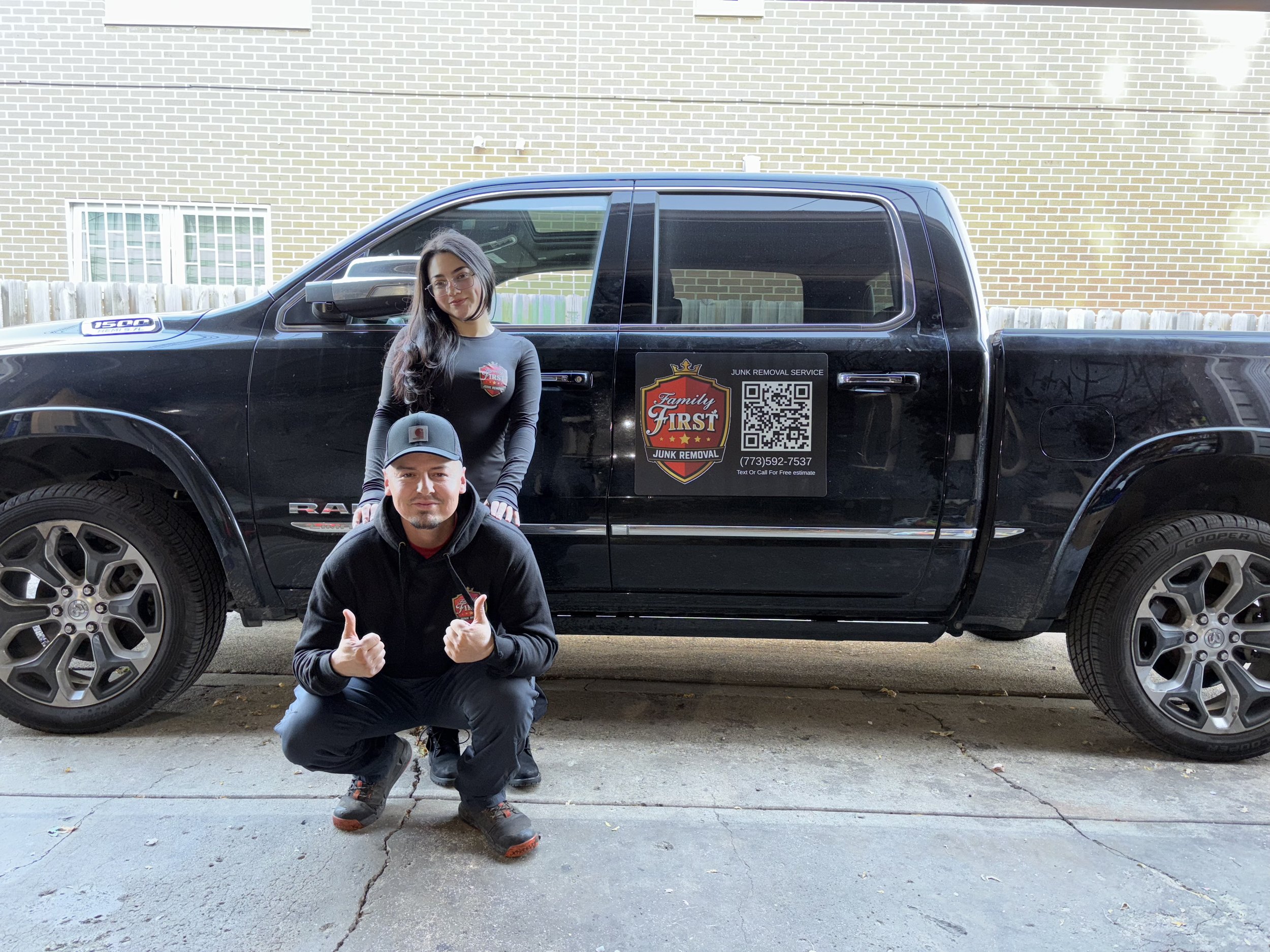 A man crouching with two thumbs up in front of a black pickup truck, with a woman standing behind him. The truck has a large logo on the side with a QR code, contact number, and company name for junk removal services.