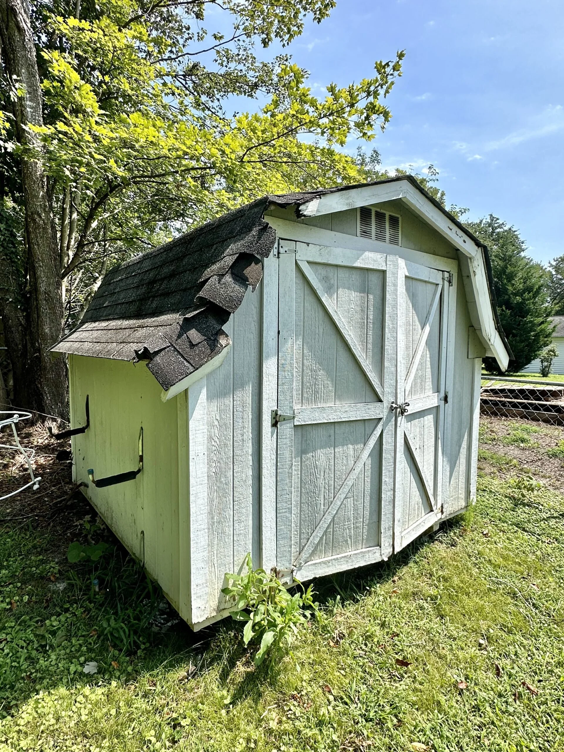 A small, weathered wooden shed with white painted walls and a shingled roof, situated outdoors on a grassy area with trees in the background.