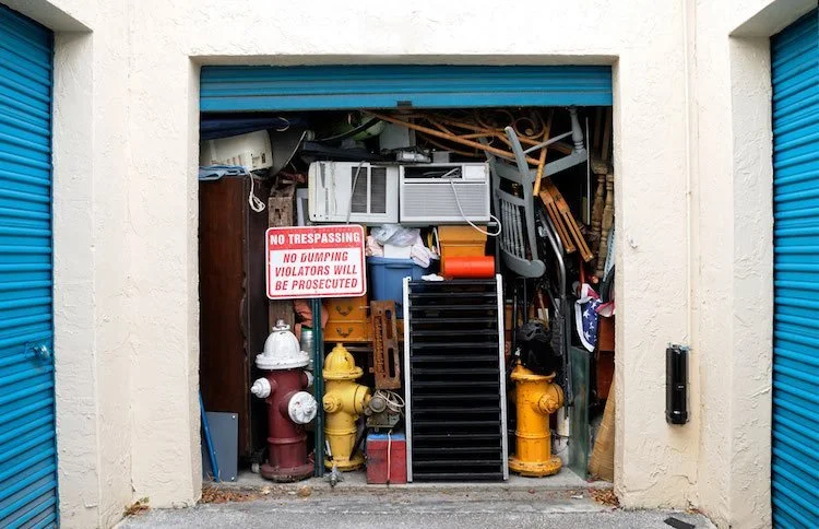 Cluttered storage unit filled with various items, including fire hydrants, a sign reading 'No Trespassing' and 'No Dumping Violators Will Be Prosecuted,' an air conditioner, wooden chairs, a ladder, and miscellaneous boxes and tools.