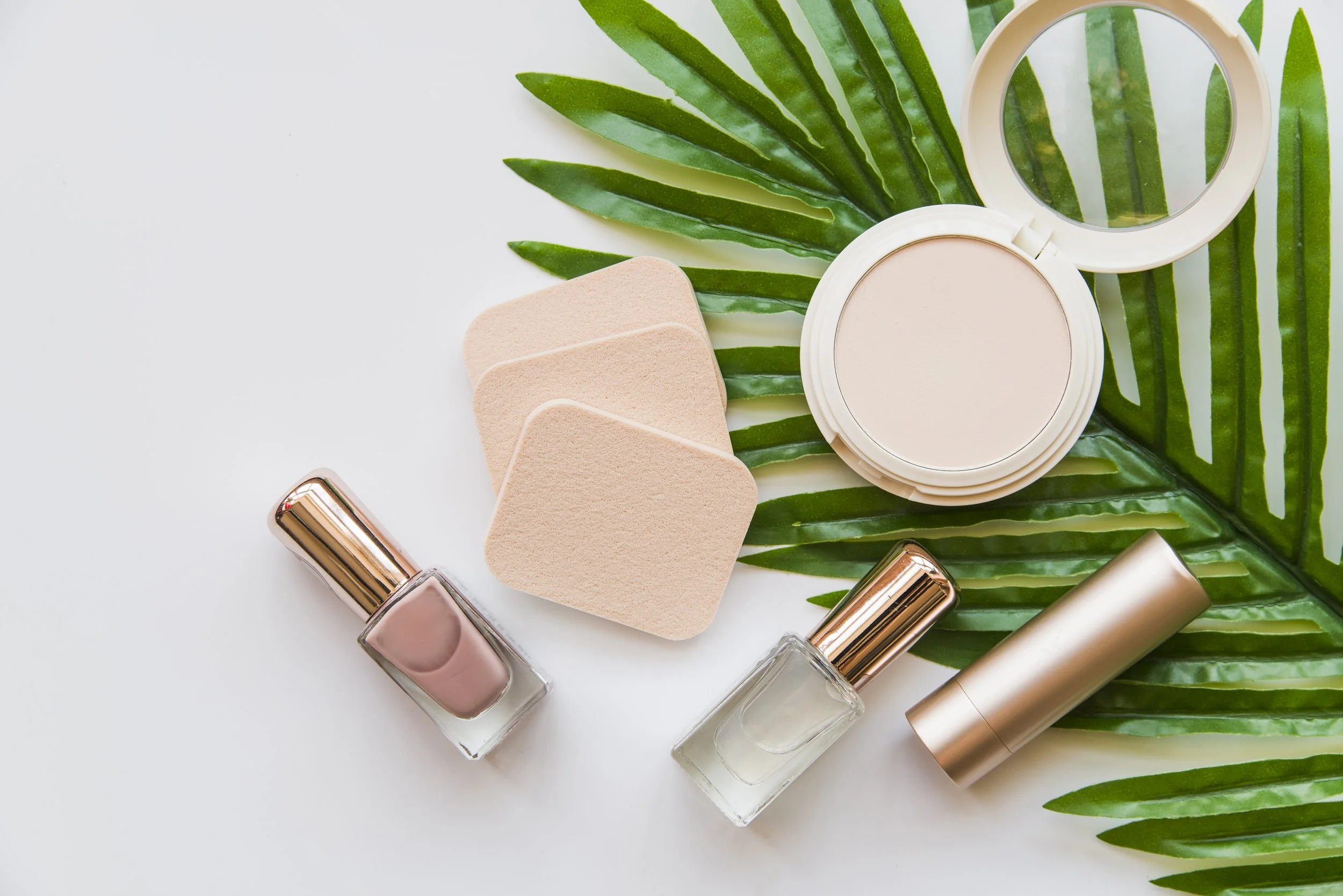 Beauty products including compact powder, nail polish, perfume bottles, a lipstick, makeup sponges, and a green tropical leaf on a white background.